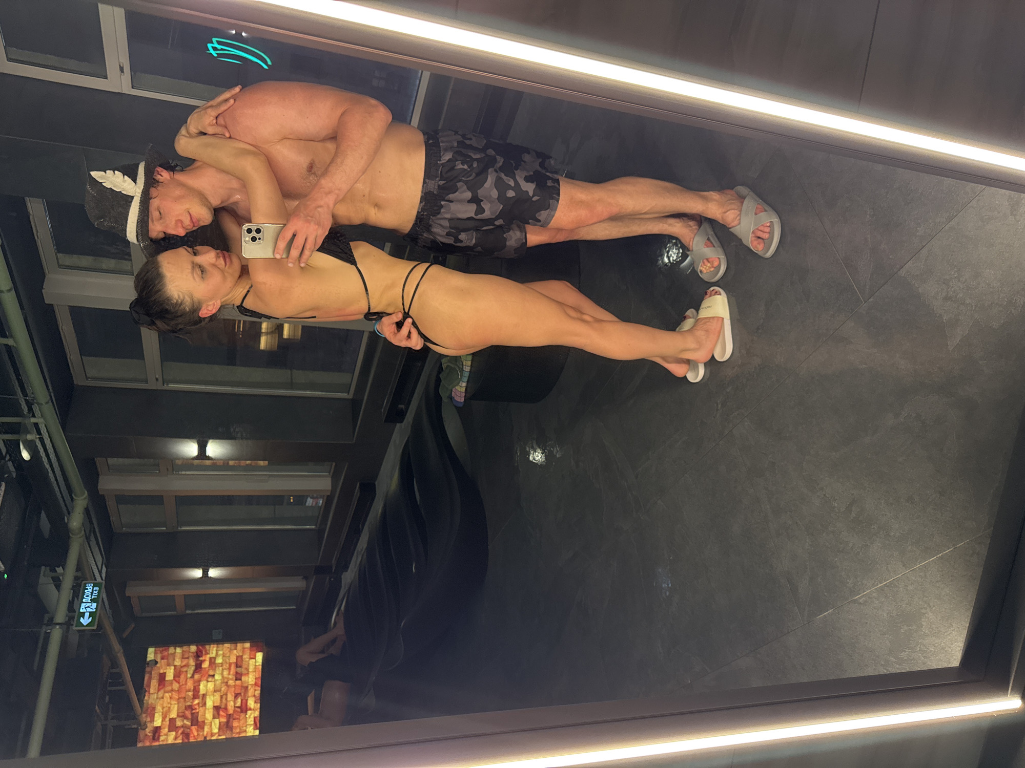 A couple stands in a sauna or bathroom, posing for a selfie in a mirror. The woman wears a black bikini and white sandals, holding a smartphone with a gold case. The man wears a traditional sauna hat, camouflage swim trunks, and gray sandals. They are embracing. The environment includes dark tile flooring, a black ceiling, and reflective walls. Outside the room, visible through a window, are buildings and a sign indicating an exit in Russian. The location is likely Saint Petersburg, Russia, based on the Cyrillic exit sign and the architectural style.
