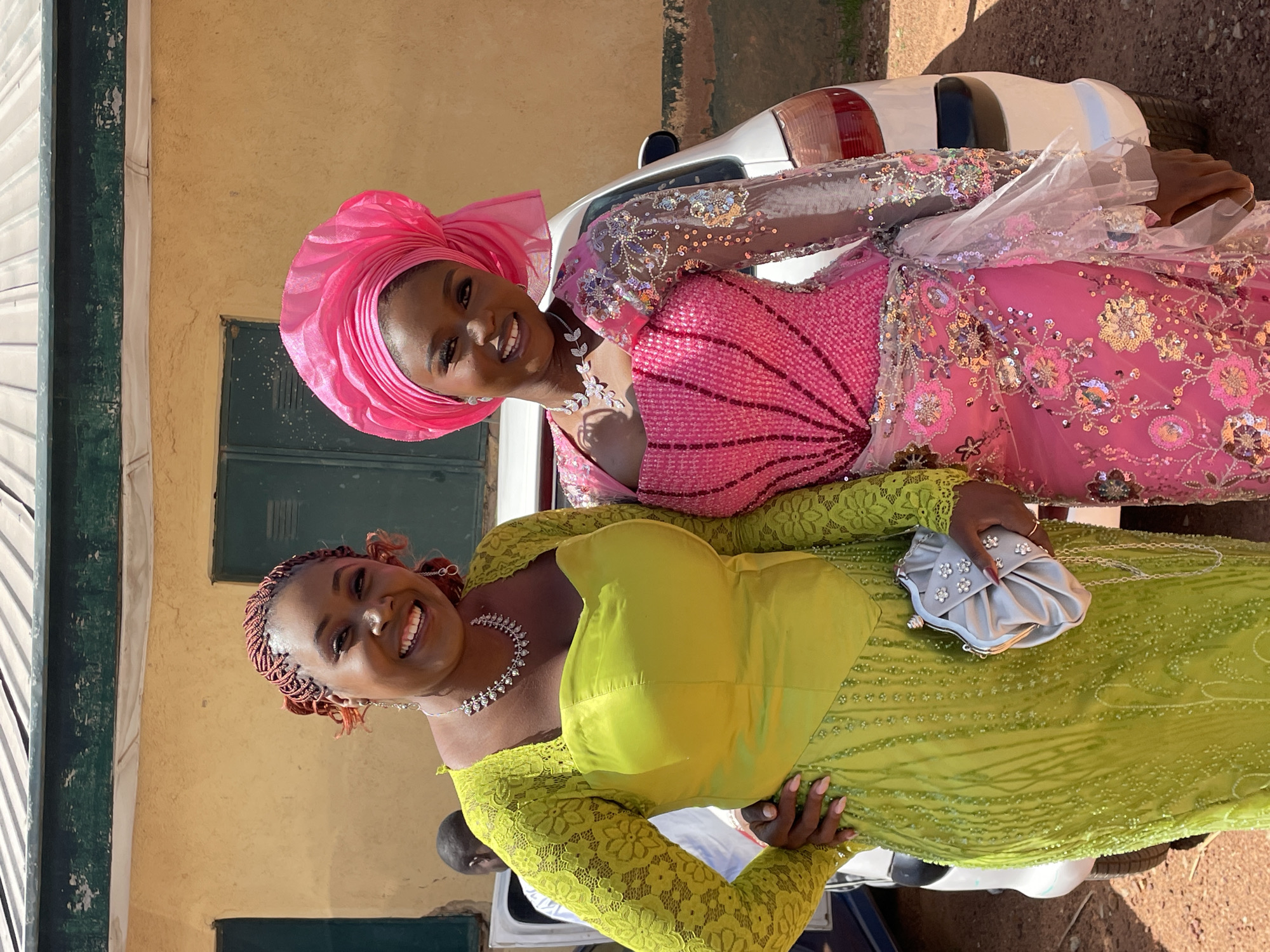 Two women are posing for a photo in front of a cream-colored building with a dark green shuttered window and a white car in the city of Jos, Nigeria. The woman on top is wearing a pink sequined dress with sheer sleeves and a pink headwrap. The other woman is wearing a yellow lace dress and holding a silver clutch. Both women are smiling and wearing necklaces.