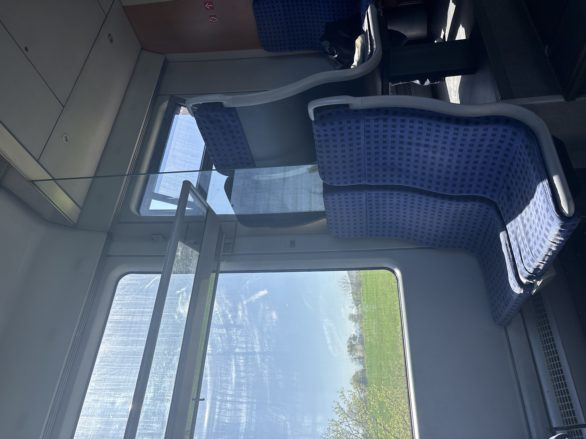 The image depicts the interior of a train car, likely during daytime. The focus is on seating and the view through the window. Blue patterned seats are visible on the right side of the image. The window showcases a rural landscape with a green field and trees under a blue sky. The window appears scratched or smudged, obscuring the view slightly. The scene does not depict Ovchepoltsi, Bulgaria, or provide any information related to that location.
