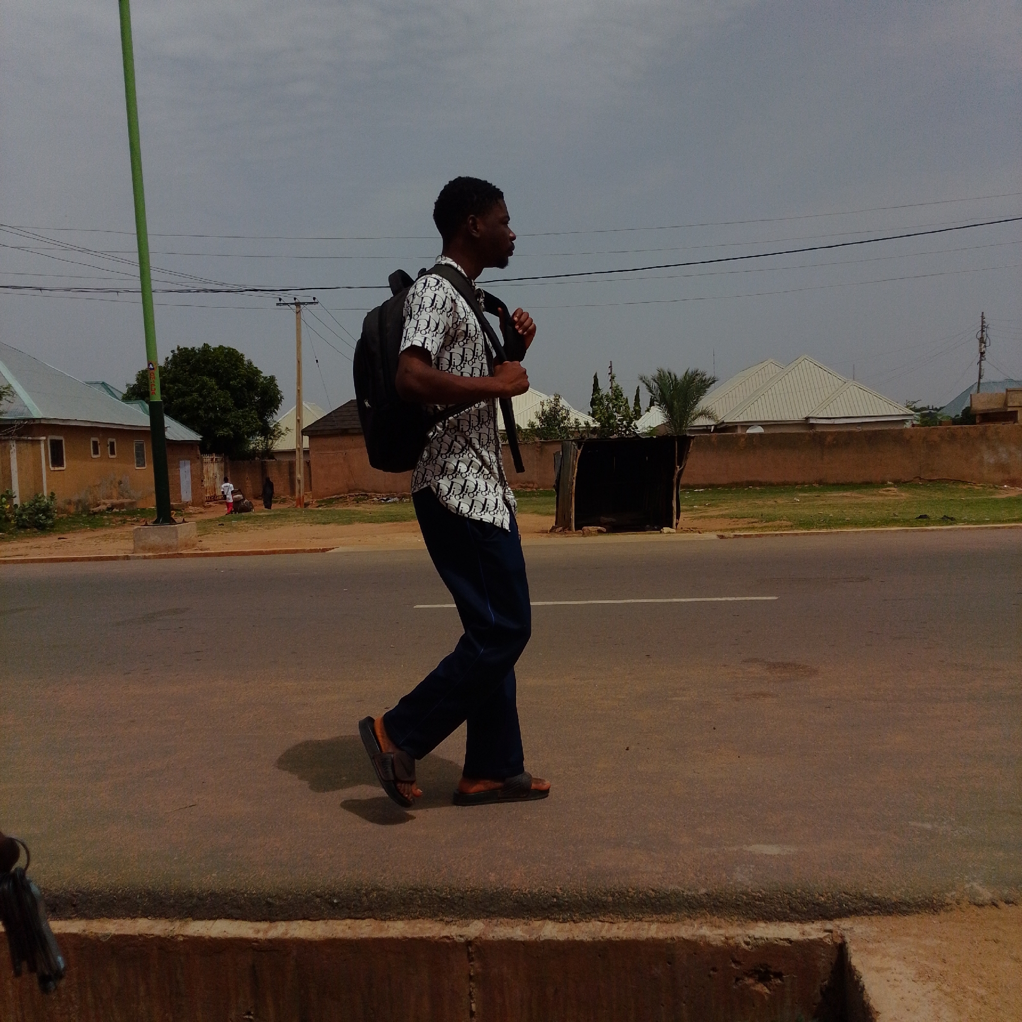 In Gombe, Nigeria, a young man walks along a paved road. He is wearing a short-sleeved shirt with a repeating pattern of "Dior" logos, dark blue trousers, and brown flip-flops. He carries a black backpack. The man is walking from left to right in the frame, his body angled slightly towards the camera. He appears to be alone.
The environment is semi-urban. To the left of the road are several single-story buildings with reddish-brown walls and light-colored roofs. There is a green pole on the left side of the image, which appears to be a street light or a utility pole. Further back, behind a low wall, are more buildings with white roofs, and some vegetation, including trees and palm trees. In the middle ground, a simple, open-sided shelter with a corrugated metal roof is visible. Power lines are visible across the sky. The sky is overcast. The foreground shows the edge of the road and a concrete structure.