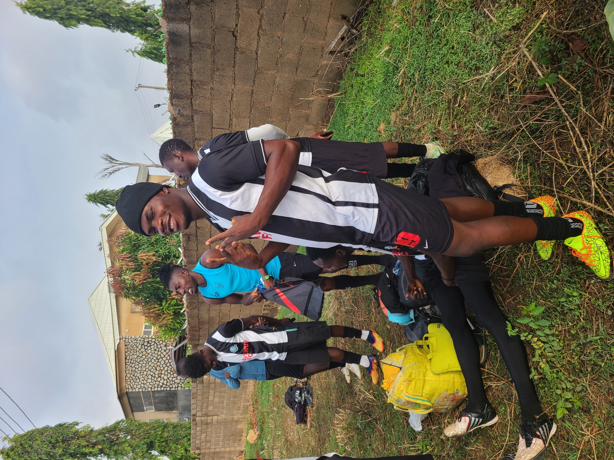 The image shows a group of young men gathered in an outdoor area, likely in Bukuru, Nigeria. The men are of African descent. Some are wearing athletic clothing, including matching black and white striped jerseys and soccer cleats. One man is wearing a blue athletic tank top. They appear to be interacting and smiling. The ground is covered with grass and some foliage. In the background is a brick wall, a building, and some trees. There are various bags and personal belongings scattered on the ground near the group.