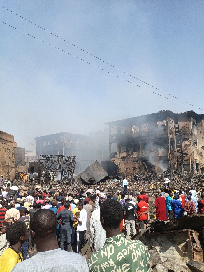 The scene in Fagge C, Nigeria, depicts the aftermath of a significant structural fire. Multiple multi-story buildings in the background are severely damaged, displaying extensive charring, collapsed roofs, missing window frames, and exposed internal structures. Residual smoke emanates from the wreckage, particularly from the building on the right. The ground in the mid-ground and foreground is covered with a dense layer of debris, including burnt materials, rubble, and twisted metal, with a large, dark, bent metal panel prominent in the mid-ground. A substantial crowd, estimated to be well over a hundred individuals, predominantly adult males, occupies the foreground and mid-ground, observing the damaged area. Some individuals are seen closer to the structures, navigating the debris. The sky above is a clear blue at the top, transitioning to a hazy light blue with smoke closer to the horizon and above the destroyed buildings, with two thin horizontal utility lines visible across the upper sky.