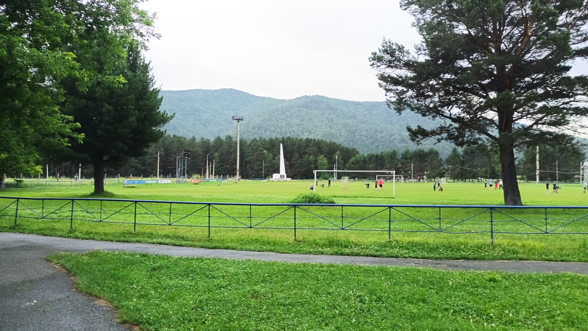 The image depicts an outdoor sports field on an overcast day. The field is a bright green expanse, likely grass, with a black asphalt path curving through the foreground. A low, blue metal fence runs along the edge of the field, separating it from the path and a grassy area.

In the middle ground, several people are engaged in various activities on the field. Some appear to be playing soccer, with a soccer goal visible on the right side of the frame and a few individuals in active poses. There are also some dogs on the field, adding to the lively atmosphere.

In the background, a white obelisk-shaped monument stands prominently, with a tall light pole nearby. Beyond the field and monument, a dense forest of evergreen trees stretches across the landscape, leading up to rolling, mist-covered mountains. The overall impression is a public park or sports complex in a natural, mountainous setting. The overcast sky suggests it might be morning or afternoon. The general mood is relaxed and recreational, with people enjoying outdoor activities.
