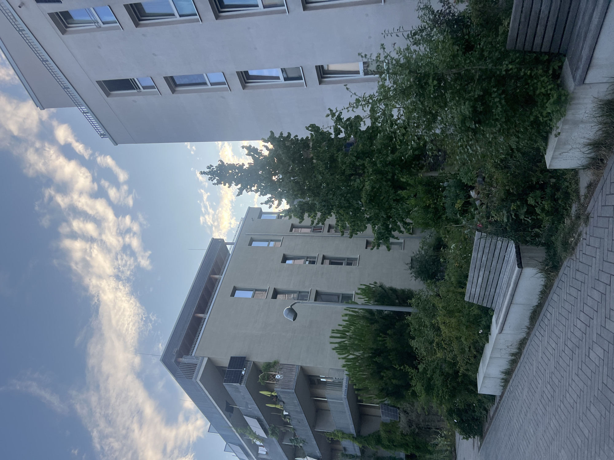 The image depicts an outdoor scene featuring two modern apartment buildings, a paved walkway, and a small park area with benches and greenery. The buildings are of contemporary design, with clean lines and large windows. The building on the left has balconies with solar panels visible. The building on the right is taller and has a lighter facade. The park area includes trees, bushes, and flowers, as well as concrete benches. The sky above is blue with scattered clouds. The lighting suggests it is either early morning or late afternoon. There are no people or animals visible in the image. The scene appears to be located in a residential area of a city.