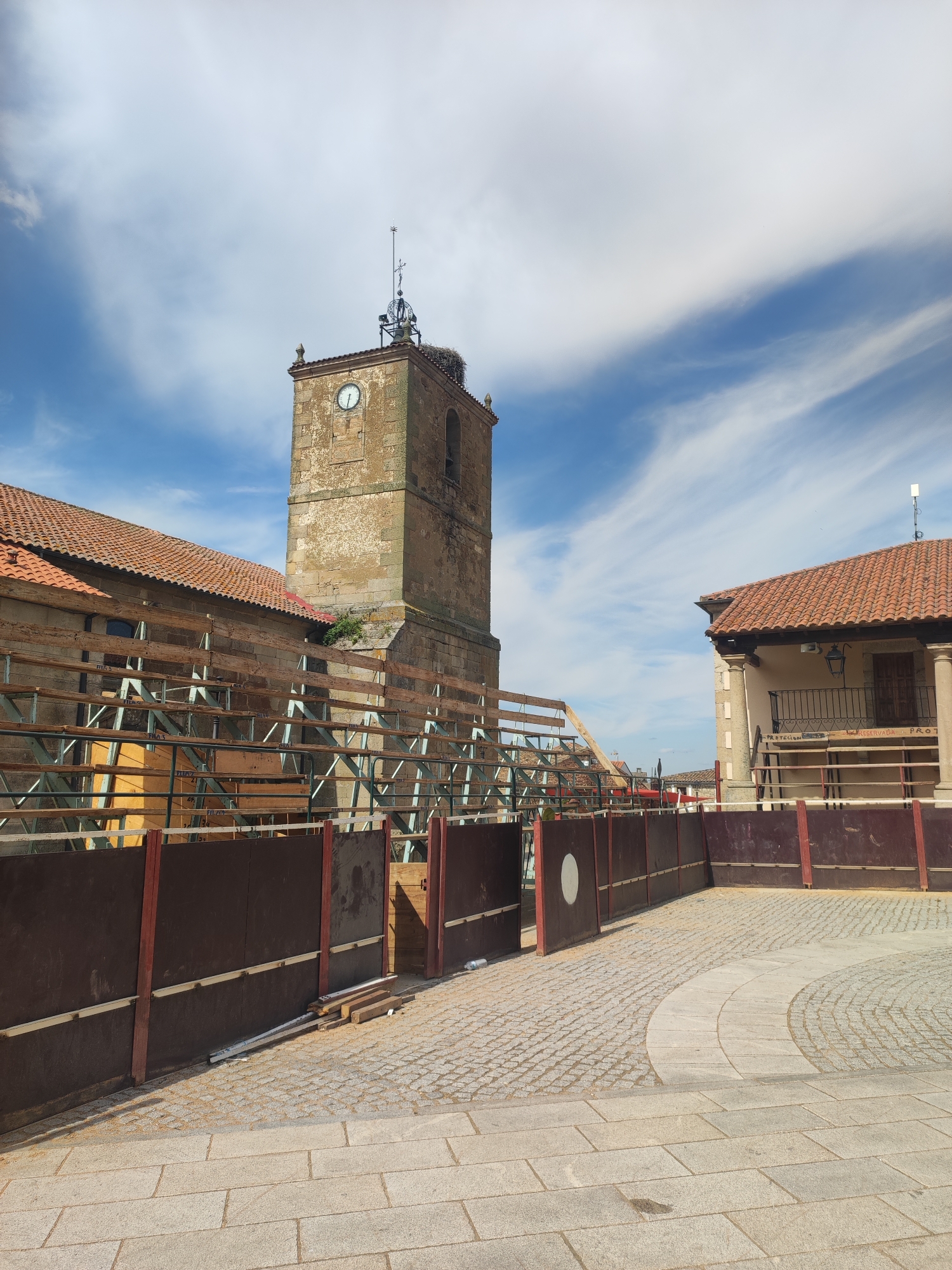 The image captures a view of the bullring in the city of Sobradillo, Spain, on a bright, cloudy day. The central focus is a tall, aged stone clock tower with a white clock face. At the top of the tower, a stork's nest is visible, perched on a metal structure. To the left of the tower, a long building with a red-tiled roof is under construction, featuring wooden scaffolding and metal supports. In the foreground, a temporary red and brown barrier encloses the bullring. The ground is paved with cobblestones and larger stone slabs. To the right, another building with a red-tiled roof has a balcony with a wrought-iron railing. The sky is blue with wispy white clouds. There are no people visible in the frame.