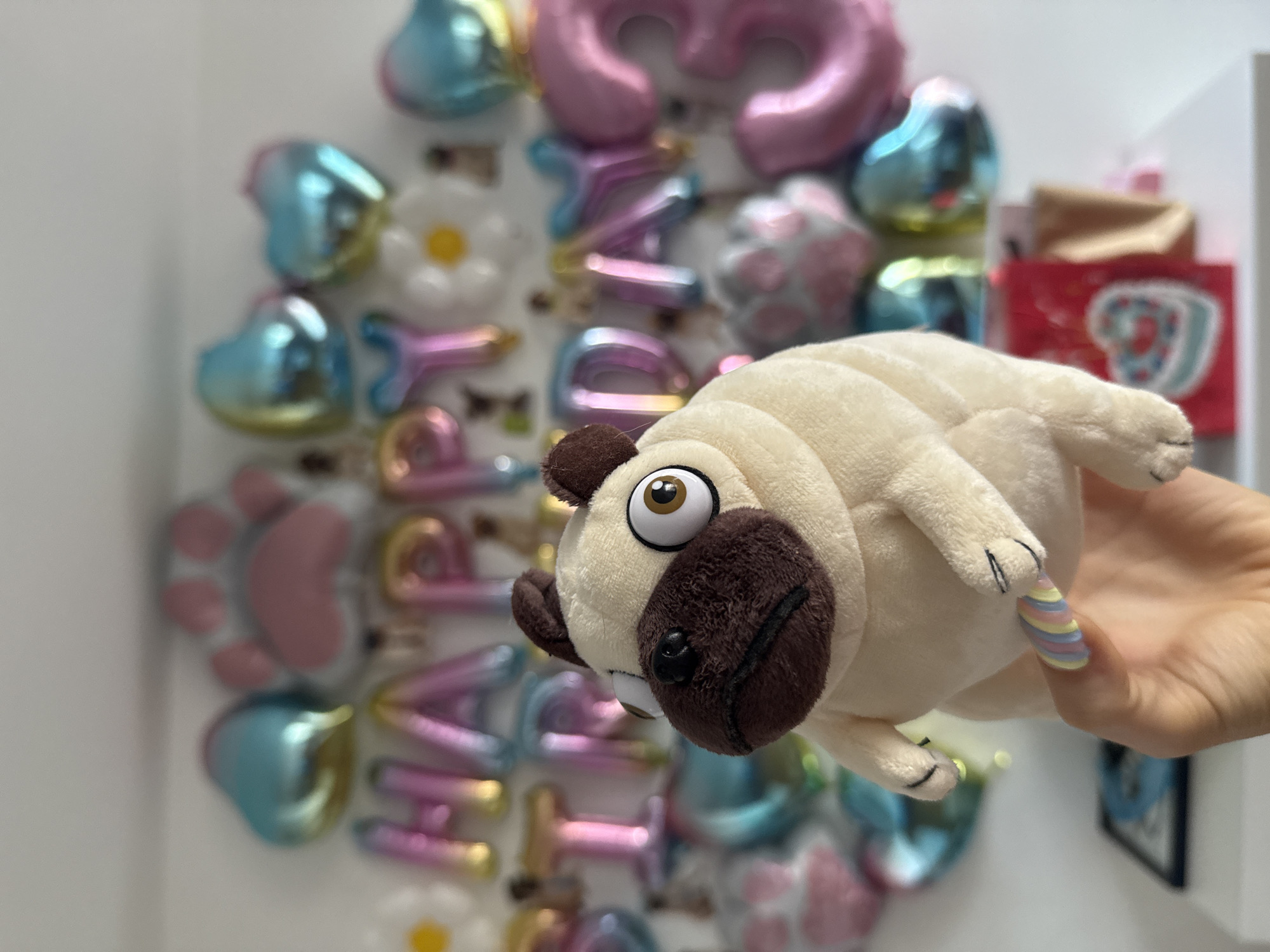 The image displays a close-up shot of a person's hand holding a plush toy in front of a festive wall decoration. The plush toy resembles a cream-colored pug, featuring a light-colored body, dark brown snout and ears, large round eyes with brown irises, and a single visible white tooth, giving it a wide-eyed, slightly surprised expression. Its paws are detailed with black stitching.
The hand holding the toy has fair skin and showcases a vibrant manicure with nails painted in pastel stripes of yellow, pink, blue, and purple.
The background is a wall adorned with numerous iridescent, metallic-finish balloons in various pastel colors such as pink, blue, gold, and purple. These balloons are shaped into letters that spell out "HAPPY BIRTHDAY," with individual letters like H, A, P, P, Y, B, I, R, T, H, D, A, and Y clearly visible. Other balloon shapes include hearts, paw prints, and white daisy flowers. A large pink number "3" balloon is also present near the top. Partially visible on the far right is a red gift bag with a light blue design, suggesting a present.
The scene takes place indoors, likely during a birthday celebration or a similar festive event, indicated by the "HAPPY BIRTHDAY" balloons and gift bag. The overall atmosphere conveyed is cheerful and celebratory. The time of day is not specifically indicated but the indoor setting is well-lit.