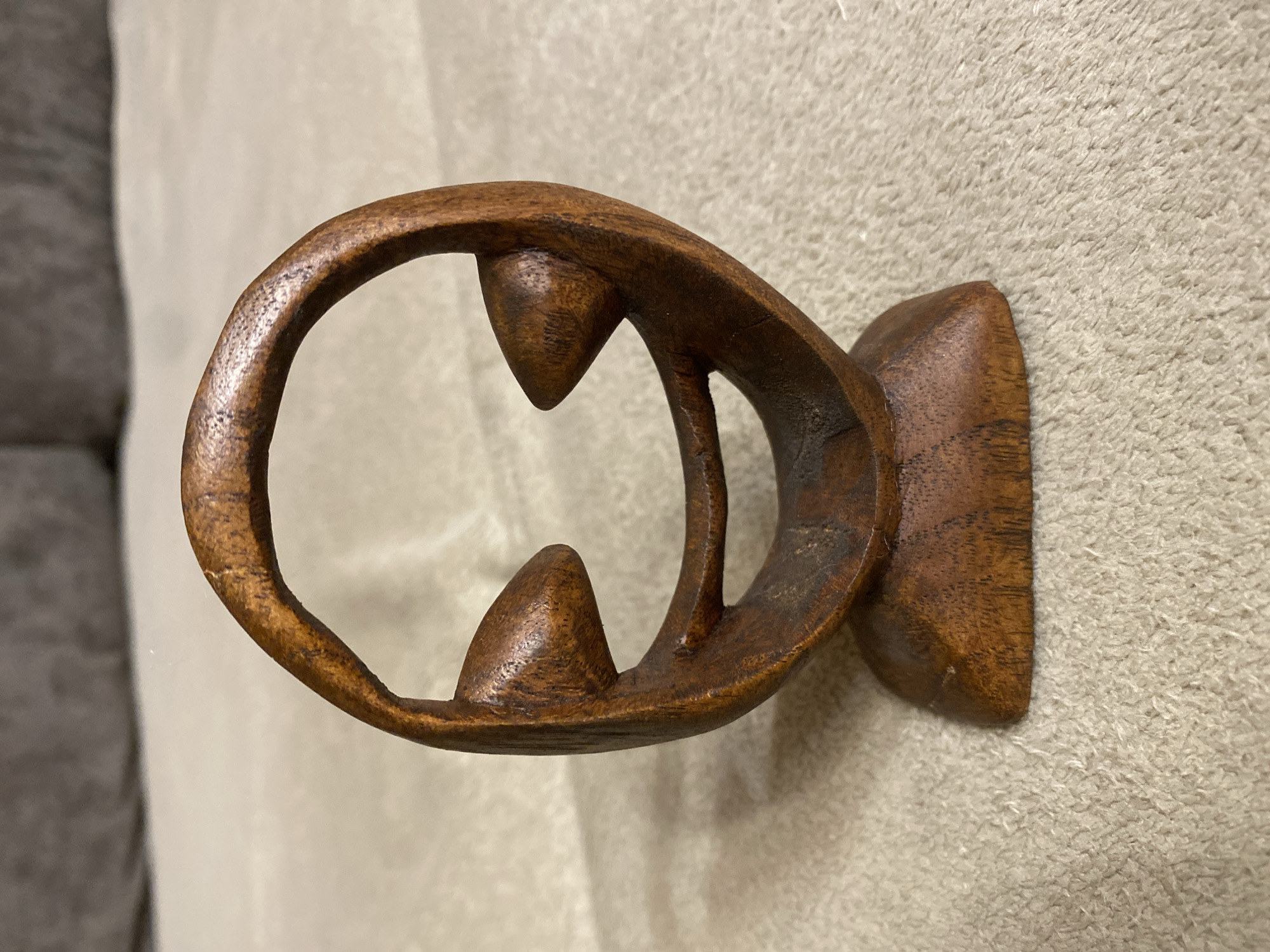 A close-up, eye-level, front-facing shot shows a small, dark brown, carved wooden figurine of a face with a circular head and two cone-shaped eyes, and a wide, curved smile. The figurine is set on a light tan, textured surface, possibly a carpet or upholstered furniture. The background is blurred, showing a darker, textured surface, likely upholstery. The lighting is even, with no harsh shadows, suggesting indoor illumination. The figurine is centrally located and in sharp focus, with the background softly blurred.