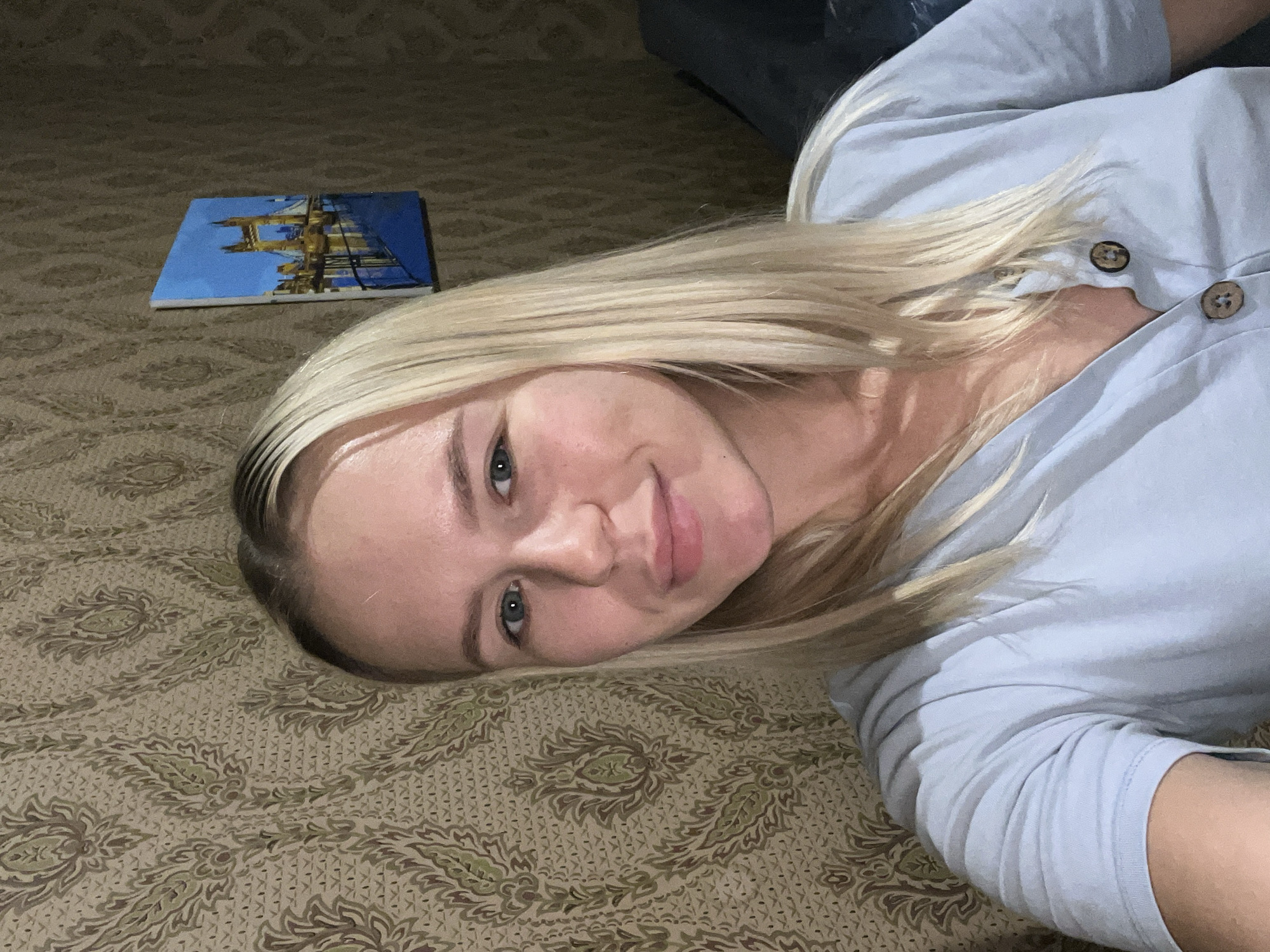 A woman with blonde hair and blue eyes is pictured in a close-up selfie taken indoors. She is wearing a light blue, short-sleeved shirt with two brown buttons. The background features patterned wallpaper with floral motifs in shades of green and brown. To the right of the woman, a small painting of the Tower Bridge in London is hanging on the wall. The lighting appears to be artificial, casting shadows on the right side of the frame.