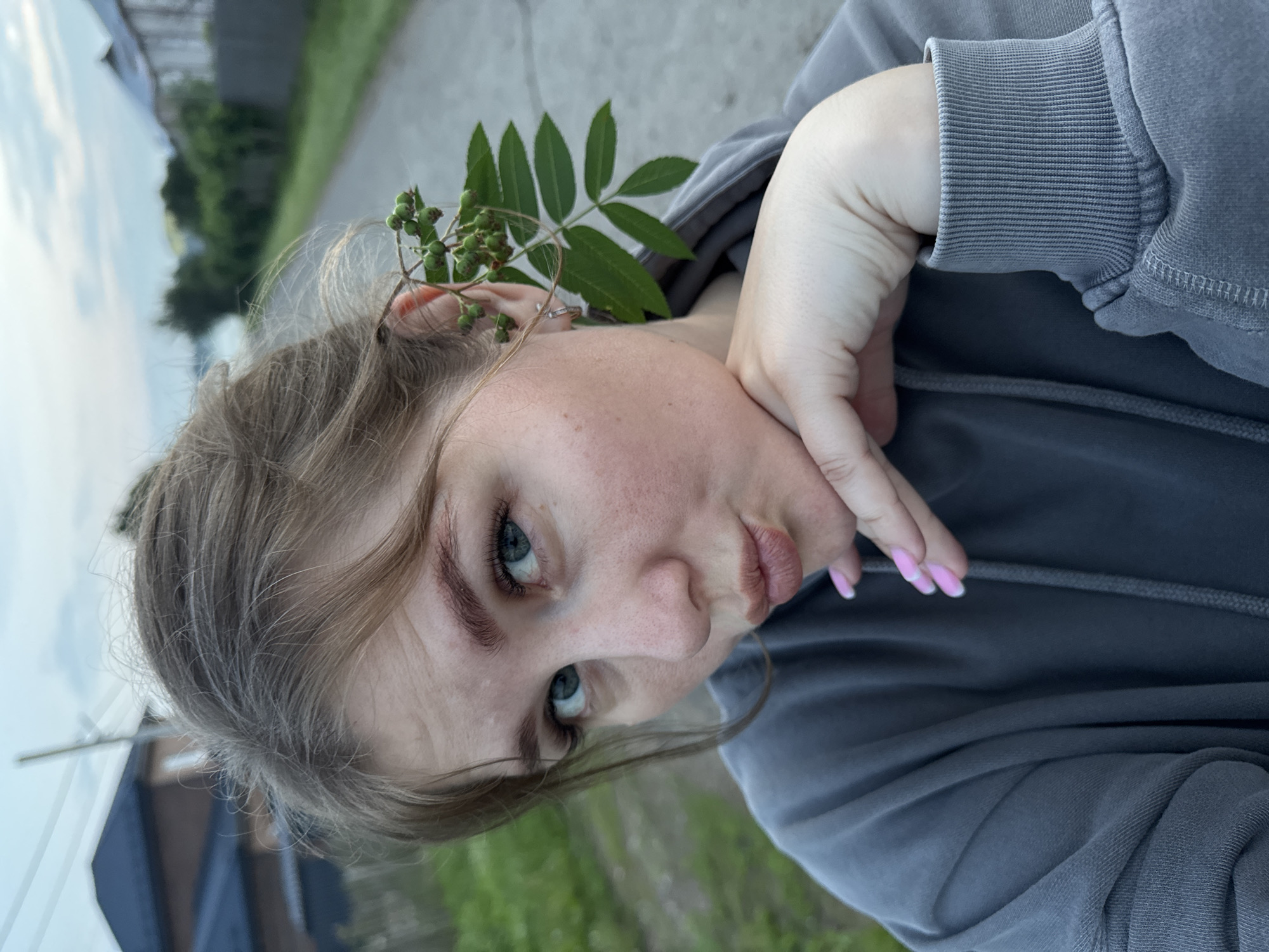 A young woman with light brown hair, casually dressed in a grey hooded sweatshirt, is captured in a close-up, outdoor selfie. She has her hand resting on her chin, and a branch with green berries is tucked behind her right ear. Her gaze is directed towards the camera, and her lips are pursed in a kissing gesture. The background suggests a semi-rural environment, with a paved path, greenery, and hints of residential buildings and a body of water, consistent with a location in the city of Gorniy, Russia. The lighting indicates it is either early morning or late evening, with a cloudy sky.