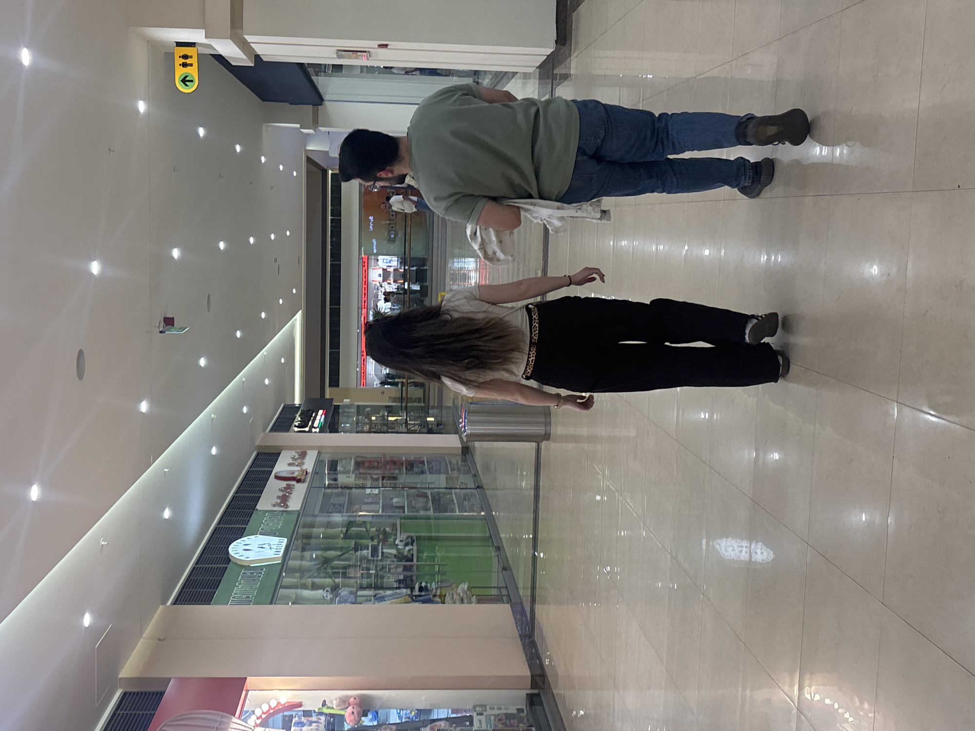 This image depicts an indoor scene within what appears to be a modern shopping mall in Yerevan, Armenia. The perspective is from behind two individuals walking away from the camera down a brightly lit corridor.
The person closer to the foreground has long, dark, wavy hair cascading down her back. She is wearing a light-colored top, dark trousers, and a patterned belt, possibly leopard print. Her left arm is slightly bent at the elbow. Behind her is another individual, wearing a dark green short-sleeved t-shirt, blue jeans, and dark shoes or boots. This person has short, dark hair and a beard, and is holding a folded white item, possibly a garment or bag, in their left hand. Both individuals are walking in the same direction, suggesting they are moving through the mall.
The environment features a highly reflective, light-tiled floor that mirrors the numerous recessed spotlights embedded in the white ceiling. On the left wall, a yellow rectangular sign is mounted, displaying a left-pointing arrow and two stick figures (male and female symbols), commonly indicating restrooms or changing facilities. The right side of the corridor is lined with glass storefronts. Partial text is visible on some shop signs: one sign behind the second person appears to show "SNA" or "SNAC" and "SWEET," while another further down features a large white clock face and possibly the text "GAME ZONE."
The bright, artificial lighting suggests the mall is open for business, likely during the day or evening. No specific emotions are discernible from the subjects as their faces are not visible. The scene captures a moment of casual movement through a commercial public space.