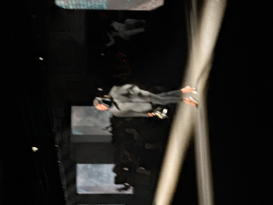 This image depicts an indoor fashion show event in Paris, France. A lone individual, presumably a model, walks purposefully on a brightly illuminated runway that diagonally bisects the frame from left to right. The individual is dressed in a dark, possibly grey, oversized jacket or top, dark trousers, and distinctive high-heeled shoes with a light-colored heel and toe. A dark hood or head covering obscures their face, and they are carrying a small, light-colored object, possibly a clutch bag, in their left hand. The surrounding environment is predominantly dark, with blurred figures of an audience visible seated in rows on either side of the runway in the background. Several rectangular screens or panels displaying faint, abstract digital patterns in blue and white hues are present in the distant background, providing additional ambient light. The scene is characterized by dramatic lighting, focusing attention on the figure on the runway against a dim, atmospheric setting.