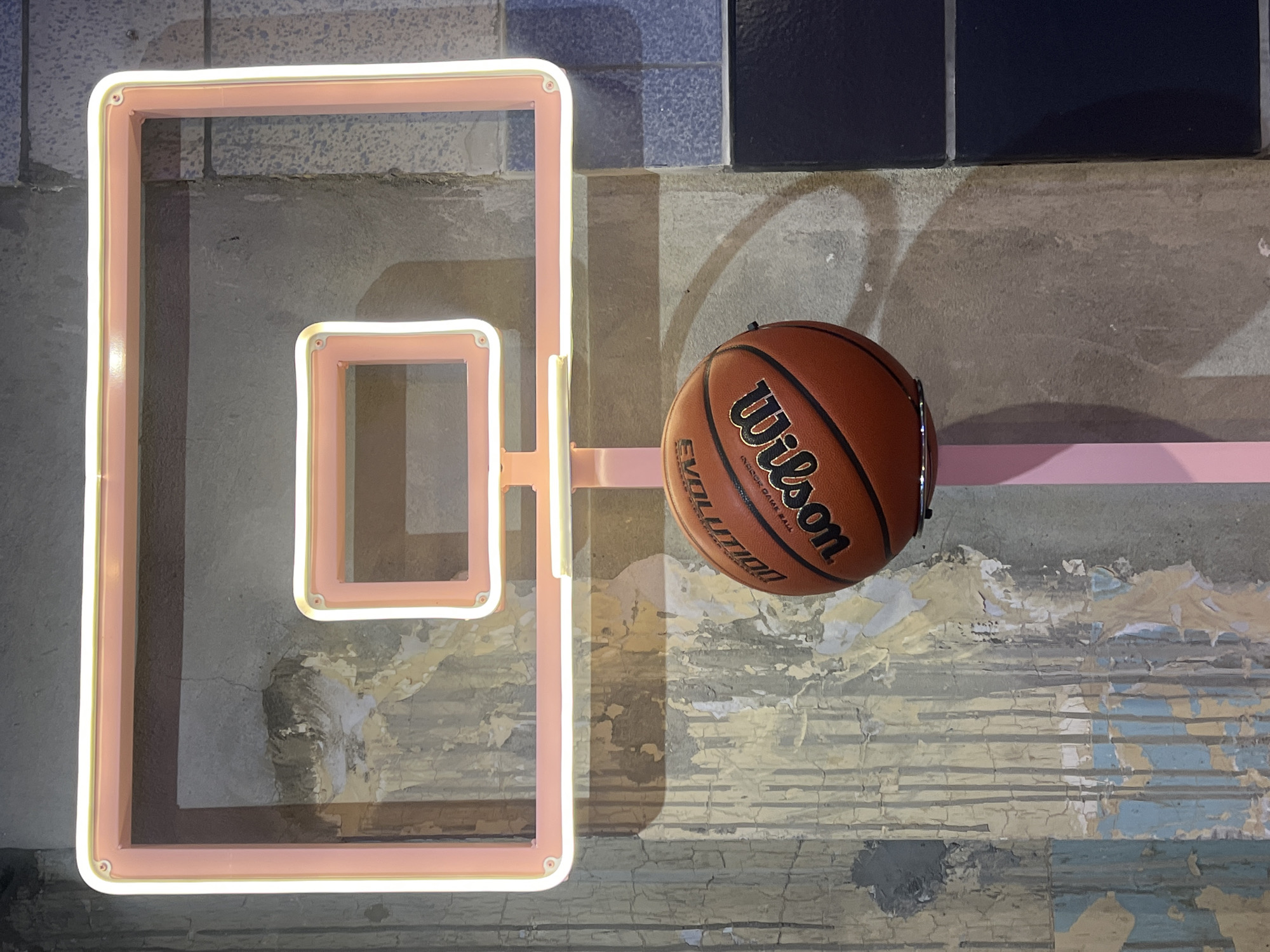 The image shows a basketball positioned on a pink structure resembling a basketball hoop. The backdrop consists of a textured wall with peeling paint and tiled sections. The structure and basketball are situated against this urban setting.
While the image provides an urban aesthetic, there isn't sufficient information to definitively determine the location as London, United Kingdom. More visual cues about architectural styles, signage, or local references would be needed to confirm its location.