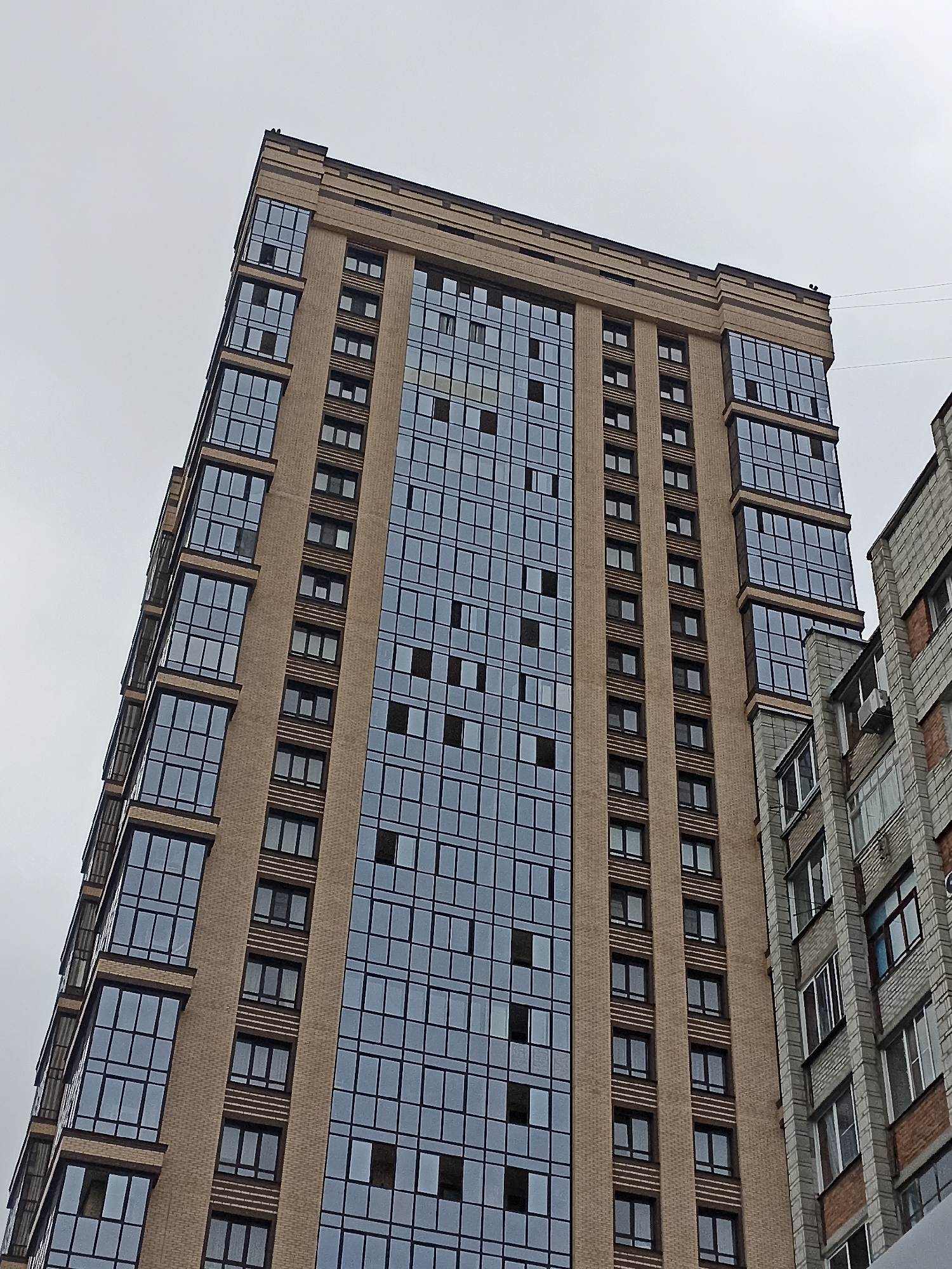 The image is an upward-angle shot of a modern, light-brown brick high-rise building with many windows, located in Novosibirsk, Russia. The building features a facade of light brown brick and large, reflective glass windows. The windows are arranged in a grid pattern, and some of them appear to be slightly darker or have the curtains drawn. The building is tall and slender, with a structured top adorned with decorative elements. To the right of the main building, a portion of an older, grey brick building with smaller windows is visible. The sky is overcast and grey, creating a muted lighting effect. There are no people or discernible activities visible in the image.