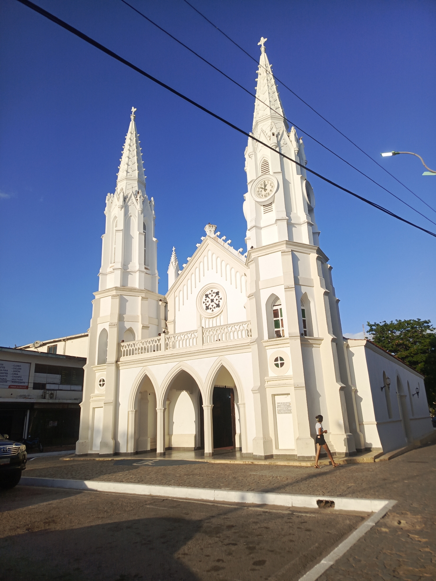 Here's a detailed description of the image:
**Scene:** The image depicts the imposing white facade of Iglesia de San Juan Evangelista, a prominent church located in Juan Griego, Venezuela.
**Architecture:** The church exhibits a neo-Gothic architectural style, with two tall, slender towers flanking the main entrance. Each tower is topped with a steeple and a cross. A clock is visible on one of the towers. The central part of the facade features arched windows, a rose window, and a balcony with a balustrade.
**People:** A woman wearing casual clothing (a top, shorts, and sandals) is walking along the sidewalk in front of the church. She appears to be looking at her phone or listening to headphones.
**Objects:**
* A car is partially visible on the left side of the image.
* Power lines run across the top of the frame.
* A street lamp is on the right side of the frame.
* A sign with text (likely advertising) is attached to the building next to the church.
**Environment:** The church is situated on a street corner. The sidewalk is paved with rectangular stones. The sky is a clear, bright blue. The lighting suggests it is daytime, with the sun casting shadows on the buildings and the street.