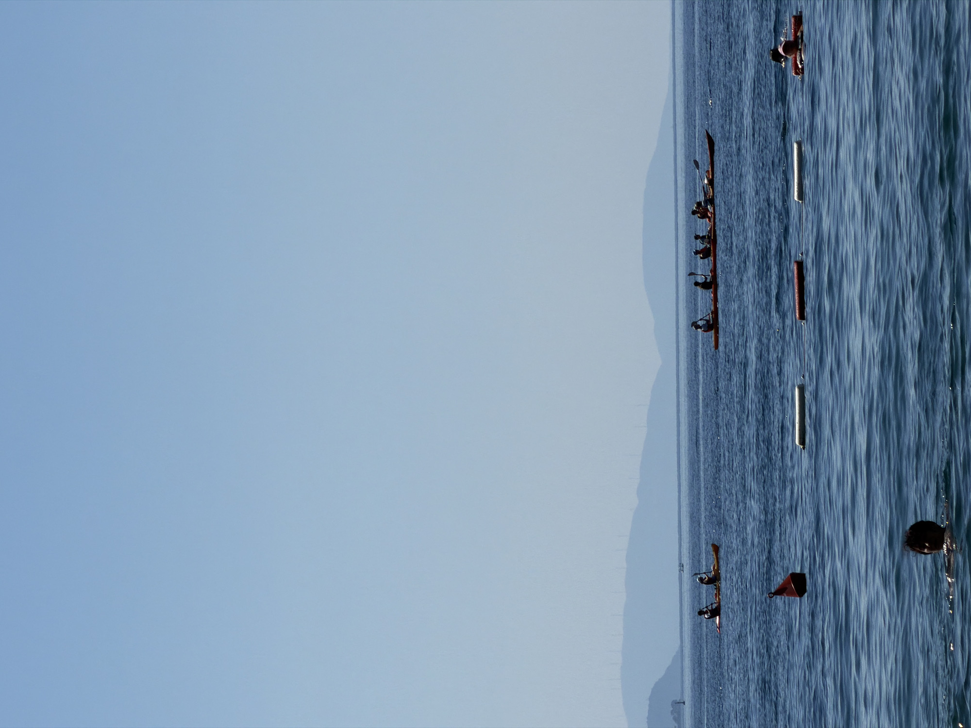 The image depicts a bright, clear day at sea with a calm water surface. In the foreground, a person's head and shoulders are visible as they swim in the water. Further out, a red buoy floats, and a white cylindrical marker lies on the water surface. In the mid-ground, a long orange kayak carries approximately six individuals, all appearing to be paddling. Behind them, another orange kayak with two people is also visible. In the far distance, hazy blue mountains form the horizon, with what appear to be wind turbines visible on their crests. The scene is consistent with a recreational area, possibly a beach or bay, offering water activities. The clear sky and calm sea suggest favorable weather conditions. Based on the visual cues, this scene could be representative of the waters off the coast of Budva, Montenegro, a popular tourist destination known for its coastline and mountainous backdrop.