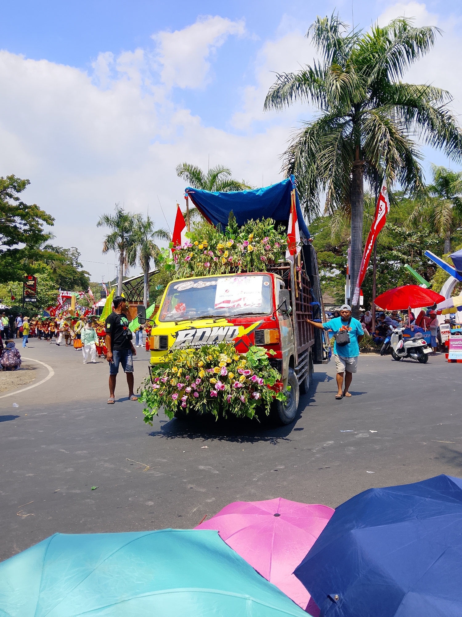The image depicts a street scene in Balun, Indonesia, likely during a parade or festival. A yellow truck, heavily decorated with flowers, is the central subject. The truck is adorned with red and white flags and a blue tarp covers part of its load. Several people are visible in the scene. A man in a black t-shirt and denim shorts stands near the truck, looking towards the left. Another man in a blue t-shirt and shorts stands to the right of the truck, near the driver's side door, with his hand on the truck. In the background, a procession of people in traditional attire can be seen walking down the street. Palm trees and other foliage line the sides of the road. Several umbrellas, in various colors including blue, pink, and teal, are visible in the foreground, suggesting a sunny day. There are also motorcycles and other vehicles parked or moving in the background. The overall atmosphere appears festive.
