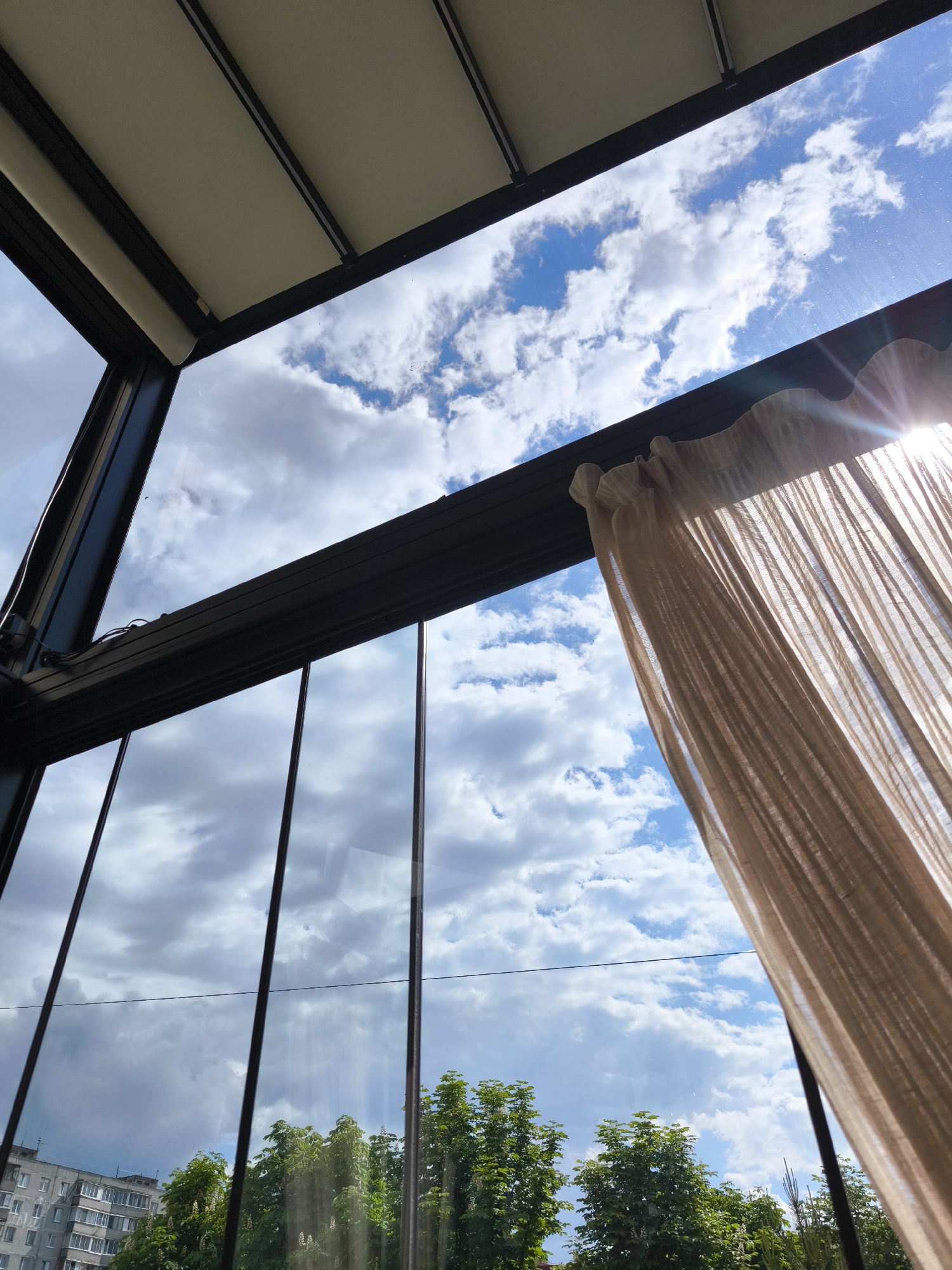 The image shows a view through a window of a structure, likely a conservatory or enclosed patio. The window is framed with dark metal bars and glass panes. In the background is a cloudy sky with patches of blue visible. A light brown curtain hangs to the right side of the window, partially obscuring the view and catching the sunlight.
Outside the window, trees are visible with green foliage. A residential building can be seen in the distance. Based on the available information (Fryazino, Russia), it's impossible to pinpoint an exact location. The image is taken from a low-angle perspective, looking upwards.