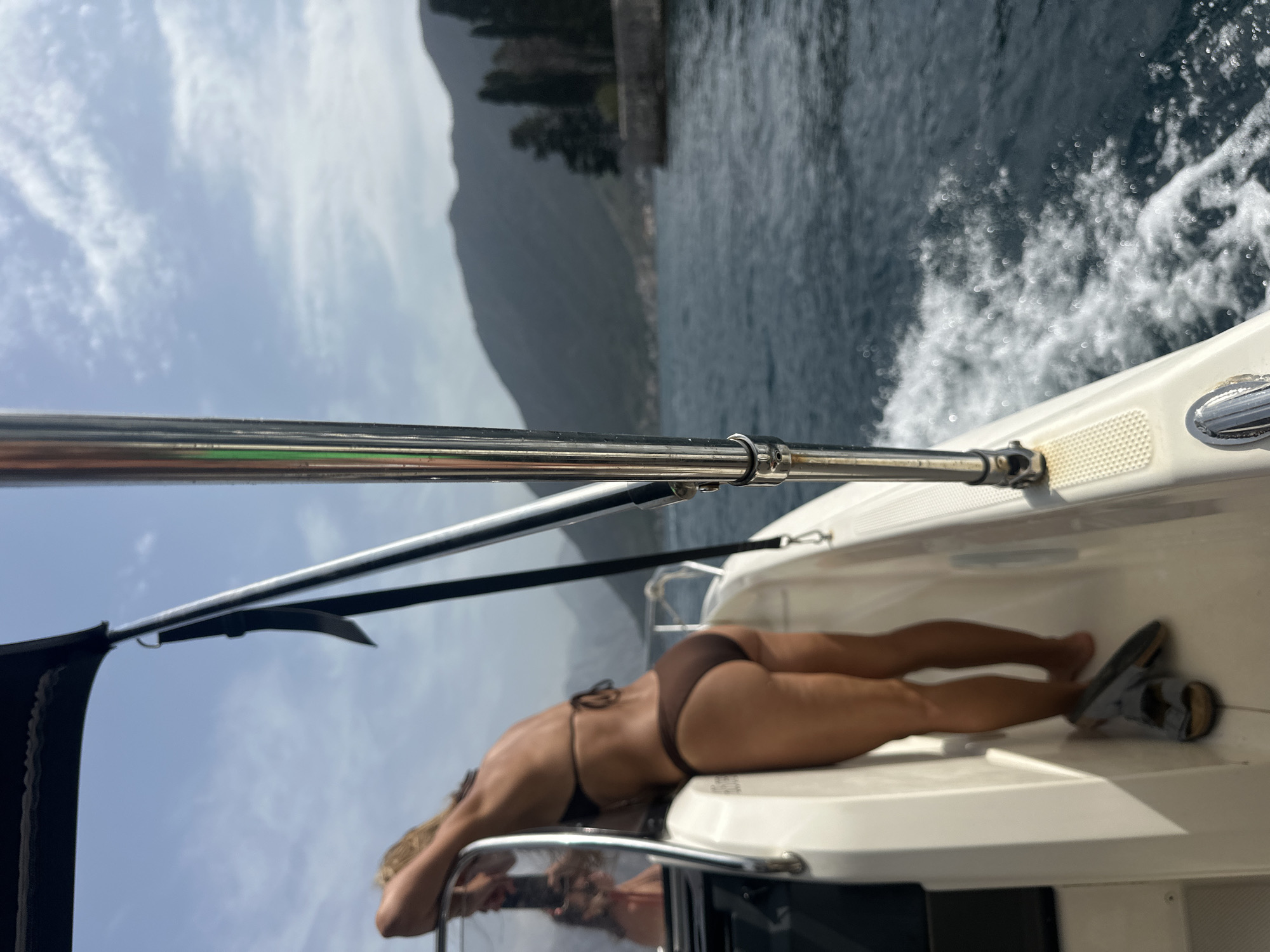 The image captures a woman in a brown bikini standing on the deck of a boat moving through the water near Perast, Montenegro. She is leaning forward, with her right arm resting on the railing of the boat. Her back is to the camera, and her legs are visible. A pair of grey sandals are on the deck near her feet. The boat is cutting through dark blue water, creating a wake. In the background, a mountainous coastline is visible, with trees and a building on a rocky outcrop. The sky is overcast with some blue patches. The perspective suggests the photo was taken from a slightly lower angle, looking up towards the woman and the boat's canopy. The chrome railing of the boat is a prominent element in the foreground, reflecting the sky and surrounding environment.