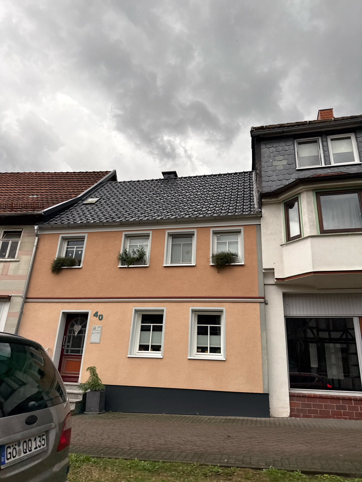 The image shows a cloudy sky over a row of buildings in Bad Sachsa, Germany. The building in the foreground is a two-story peach-colored structure with a dark gray base. It has several windows, two on the upper level with plants in window boxes, and one on the lower level to the right of the entrance. The entrance itself is a red door with a glass panel and a decorative heart shape. Above the door, the number "40" is displayed, along with a mailbox. To the left of the peach building is another building with a red tiled roof. To the right of the peach building is a building with a dark gray, slate-like roof and a white bay window on the upper level. The lower level of this building appears to be a storefront with a large dark window. A silver car with the license plate "GÖ QQ 135" is parked to the left of the peach building, partially obscuring the lower left portion of the image. Paved walkway with brick patterns is in front of the buildings, and a small patch of grass is to the far left.