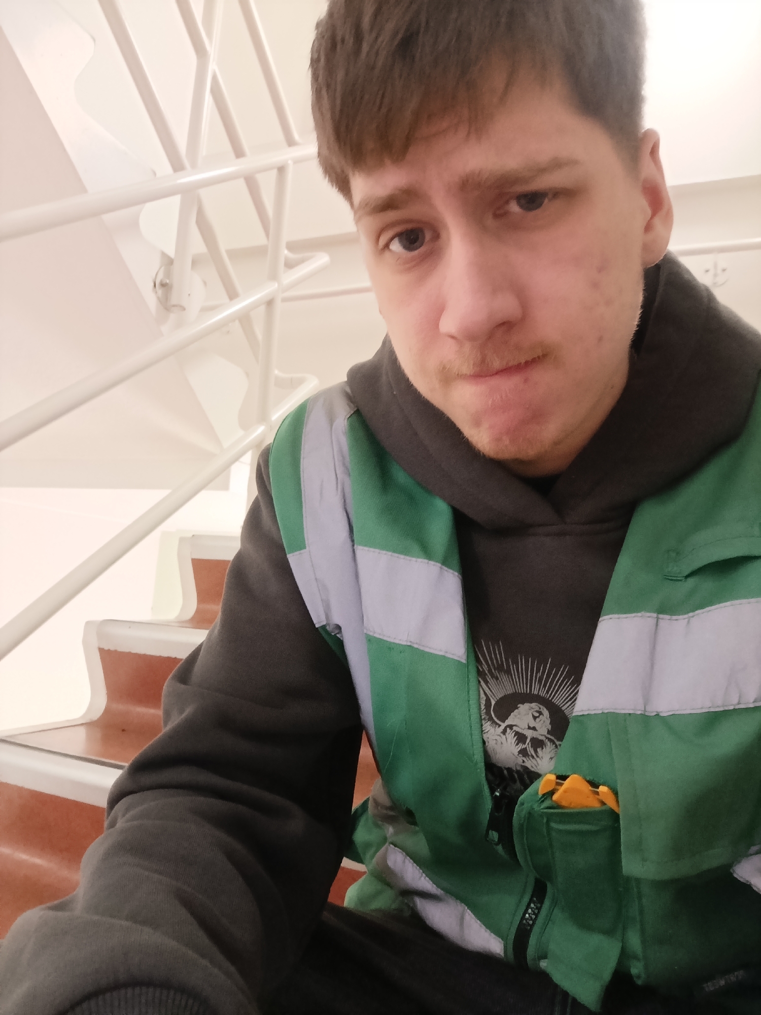 A young, fair-skinned male with short brown hair, a mustache, and visible acne, is sitting on stairs. He is wearing a grey hooded sweatshirt and a green and grey reflective safety vest. His right pocket appears to contain an orange object. The stairs have white railings and light brown treads. The location is possibly in Duiven, Netherlands.