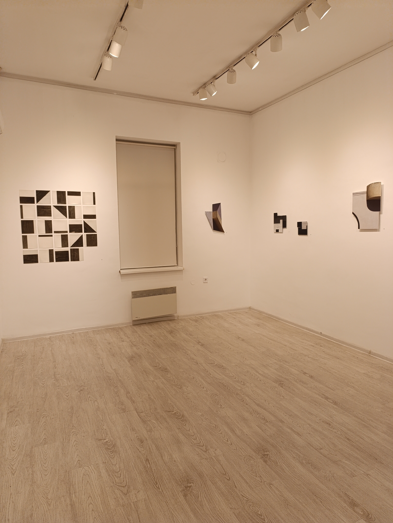 The image depicts an interior view of an art gallery in Sofia, Bulgaria. The gallery features white walls, light wood flooring, and track lighting. On the left wall, a grid-like artwork of 4x4 squares with black and white geometric patterns is displayed. Adjacent to it, a window with a closed blind is positioned above a radiator. The right wall showcases three separate abstract artworks: one with a curve and brown element, another with two black square shapes, and a third abstract piece on the far left. The gallery appears empty, with no people visible in the frame.