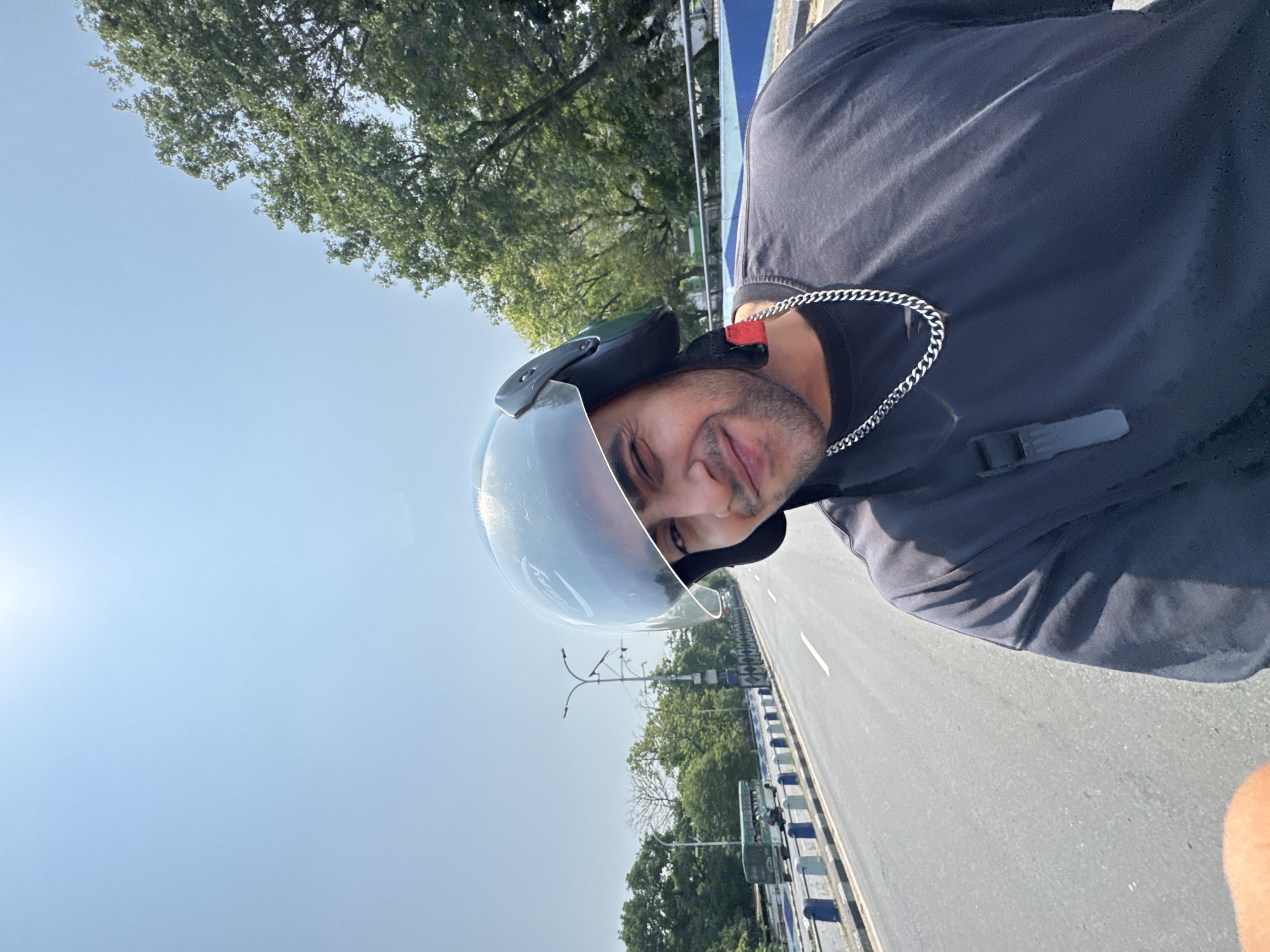 The image depicts a person wearing a motorcycle helmet with a clear visor, standing on a road. The person is wearing a black shirt. A silver chain necklace is visible. The environment is a road with a bridge. Trees line the side of the road. The sky is light blue. The location appears to be in Kolkata, India, based on the style of the bridge railings and street lighting.