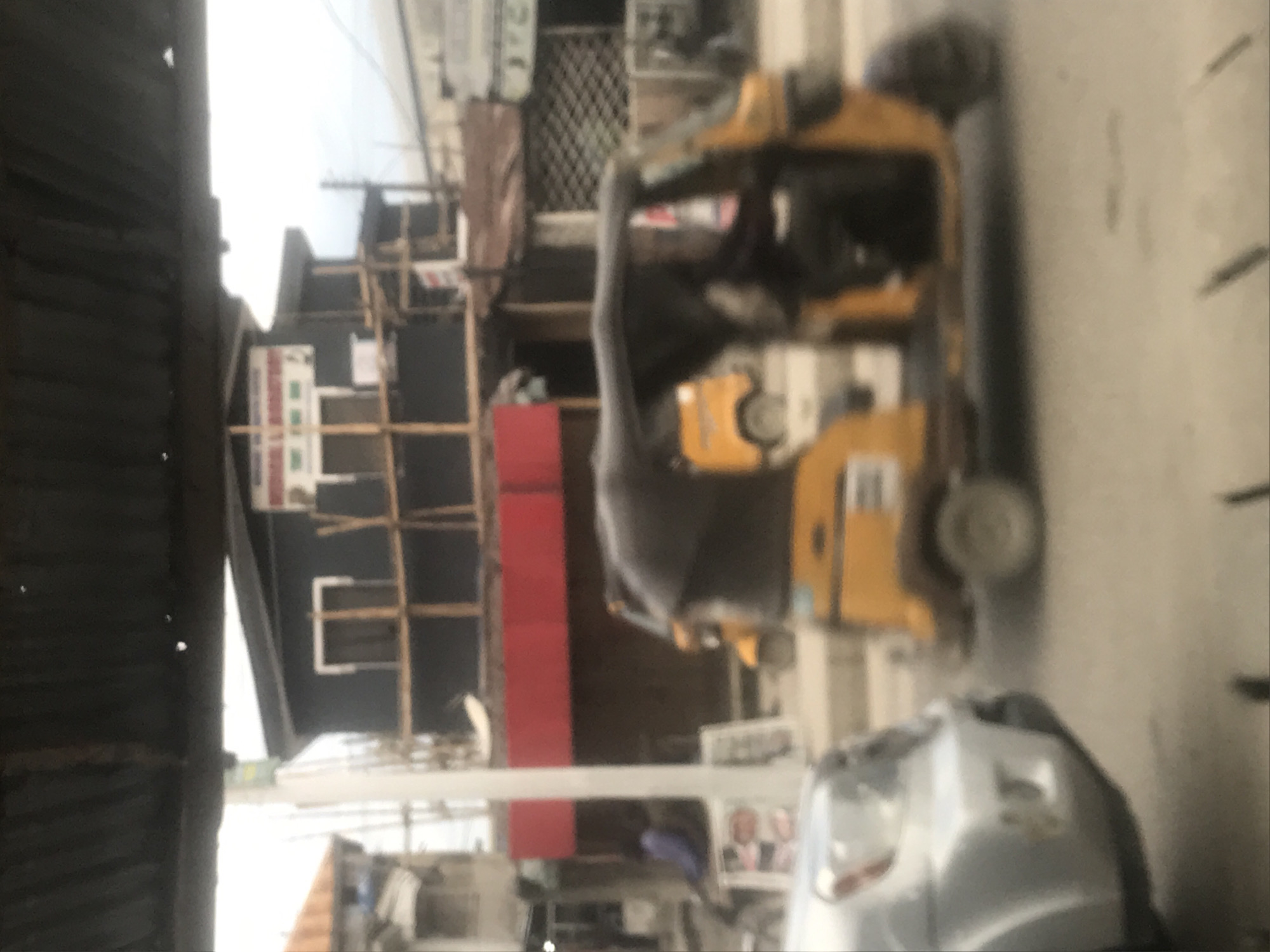 The heavily blurred and rotated image depicts an outdoor urban street scene during daytime. The most prominent subject is a yellow-orange auto-rickshaw, or 'tuk-tuk' style vehicle, positioned in the middle-right, facing towards the right side of the frame. The front of a silver or grey vehicle, possibly a car, is partially visible in the bottom-right corner.
On the left side of the image, a dark-colored structure, possibly a wall or building made of corrugated material, occupies a significant portion of the foreground. In the background, behind the auto-rickshaw, several buildings are visible. One building features a black facade with what appears to be light-colored scaffolding or structural elements. Below this, a section of a building with a solid red wall is present. The ground beneath the vehicles is a light-colored, likely paved road surface.
The overall scene suggests a typical street environment in an urban or semi-urban area. The lighting indicates it is daytime, with bright but diffused light, possibly under an overcast sky. The style of the auto-rickshaw is a strong location cue, commonly found in South Asian and Southeast Asian countries. Due to the extreme blurriness of the entire image, no specific activities of people or vehicles are discernible beyond their presence on the street. No text on any signs, vehicles, or buildings is legible.