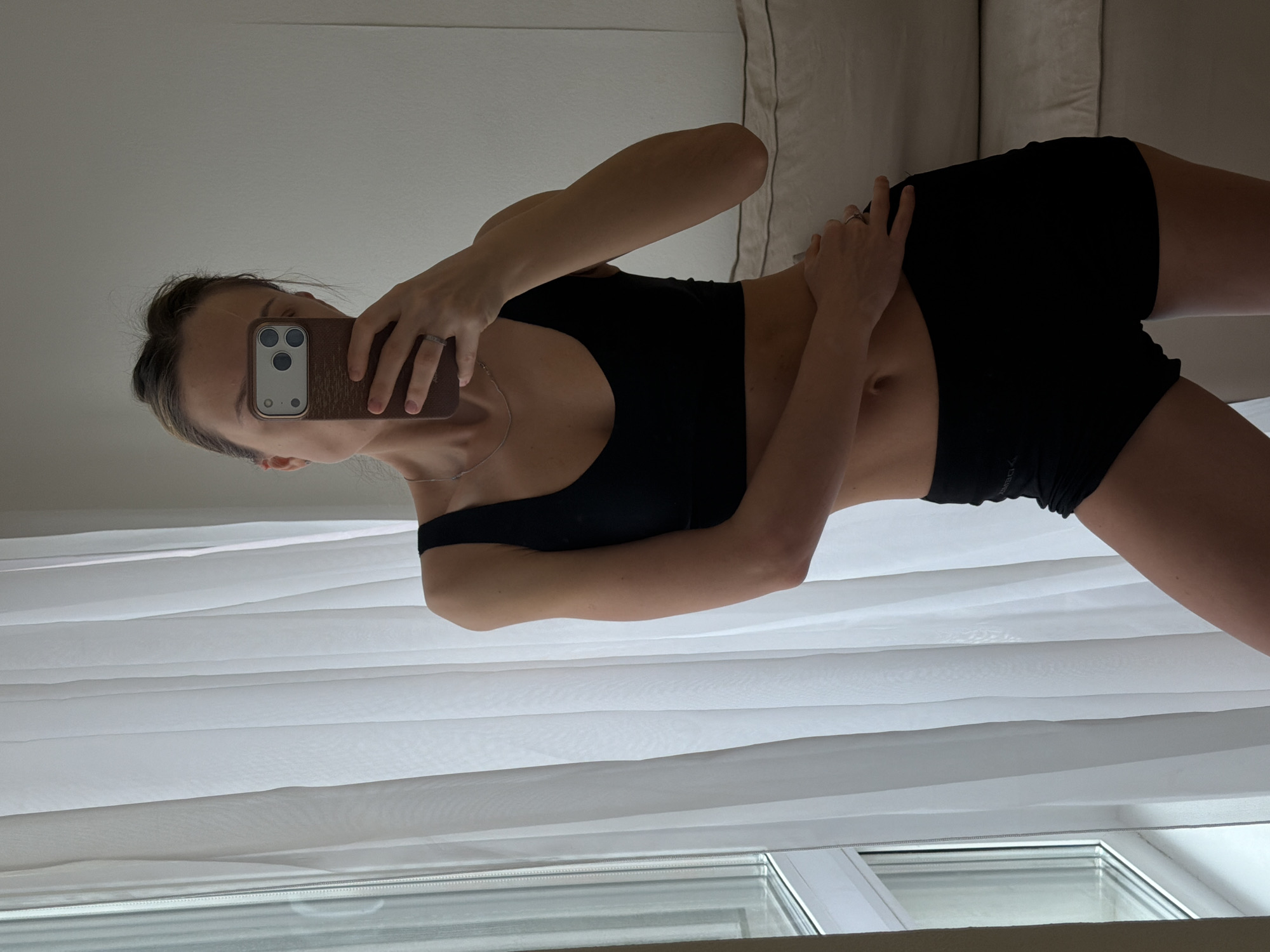 A young woman stands in front of a mirror, taking a selfie with her smartphone. She is wearing a black sports bra and black shorts. Behind her, there are white curtains and a window. The scene includes a partial view of a couch. The image provides no indication of the location being in Khimki, Russia.