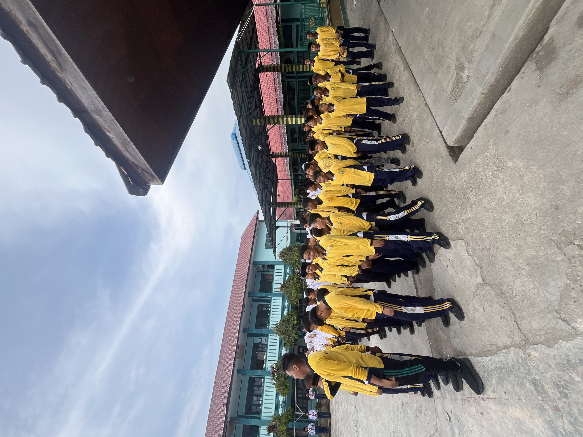 A group of male students in yellow shirts and dark blue pants stand in formation outside a teal-colored building with a red roof. The students are lined up in rows on a paved courtyard. The building appears to be a school, with windows and balconies visible. A covered walkway with yellow and green pillars runs alongside a portion of the building. The sky is overcast with scattered clouds. The scene is likely in the city of Lhokseumawe, Indonesia.
