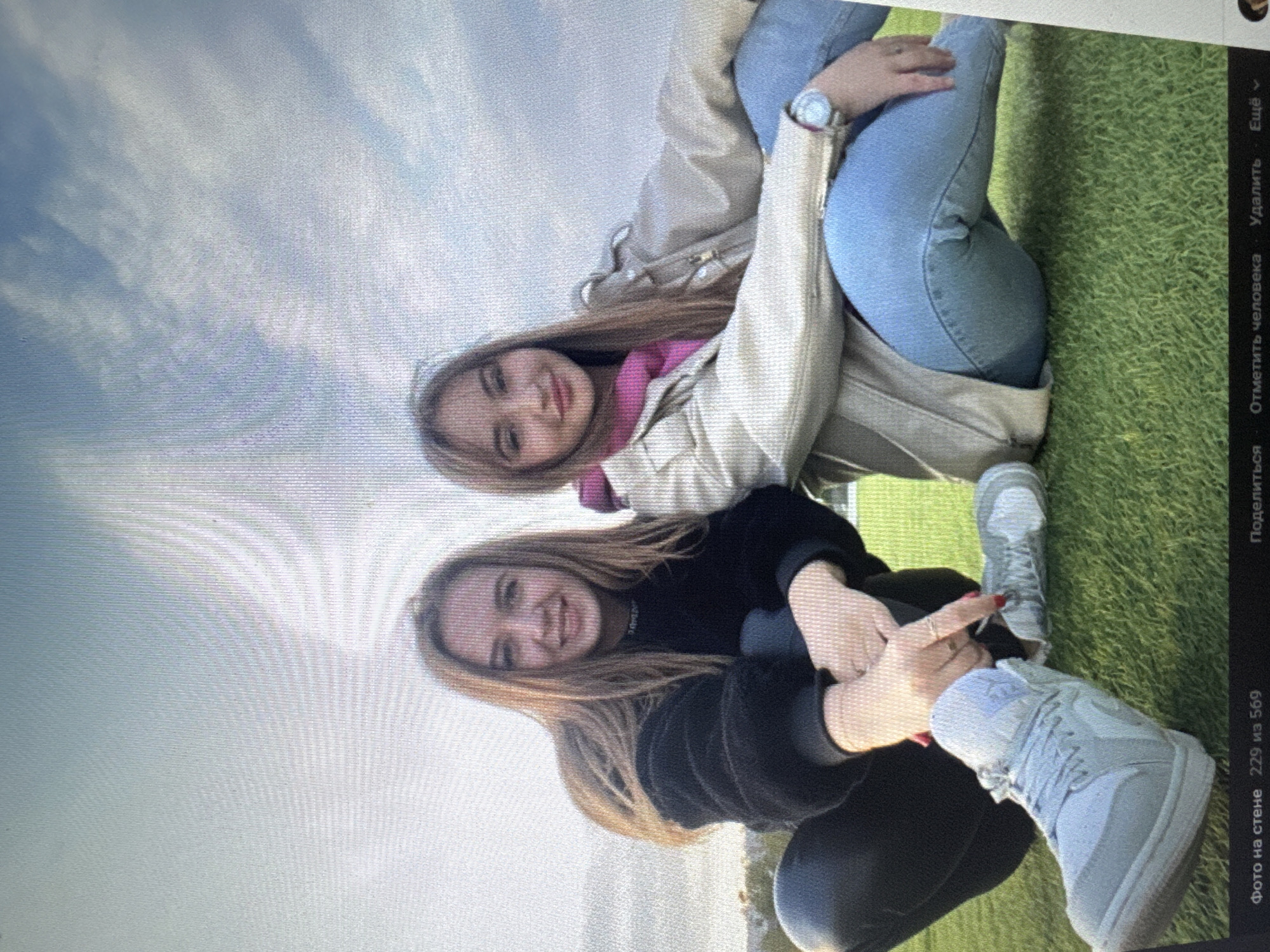 Two young women are seated on a grassy field with a cloudy sky in the background. The woman on the left has long blonde hair and is wearing a black fuzzy jacket and black pants. The woman on the right has long blonde hair and is wearing a light-colored jacket over a pink hoodie and blue jeans. Both women are looking towards the camera and smiling. They are positioned in a relaxed, seated posture, with their legs extended. The ground beneath them is covered in green grass. The overall impression is a casual outdoor portrait.