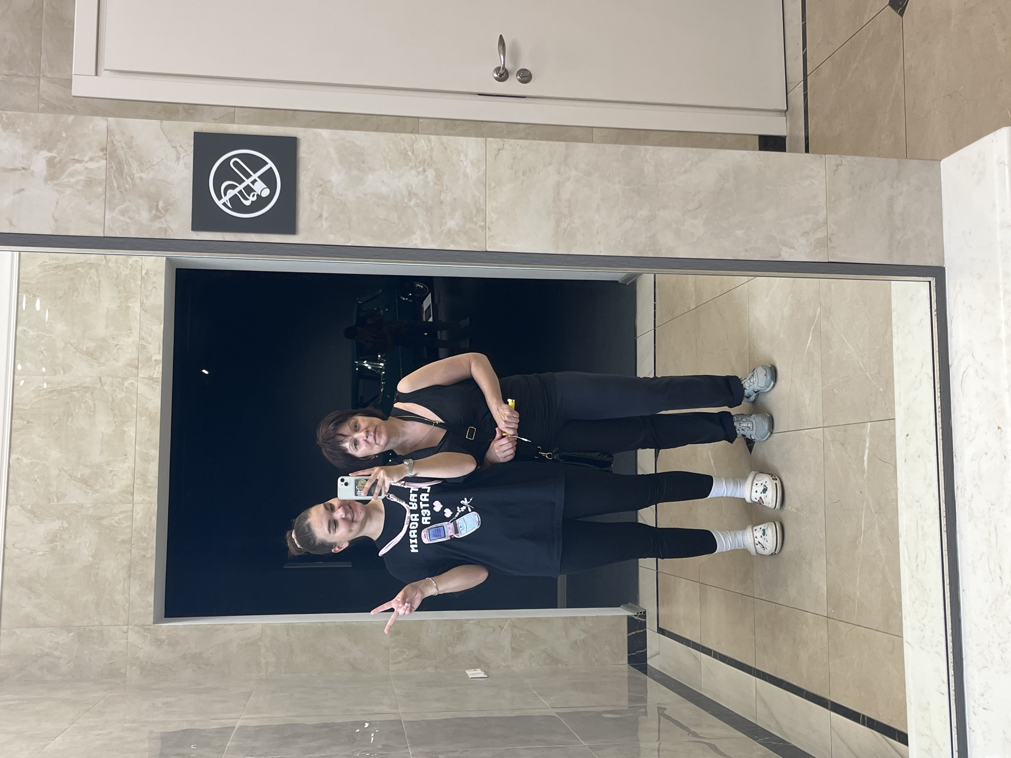 In Verkhnyaya Pyshma, Russia, two individuals, an adult female and a young female, are captured in a mirror selfie. The young female, on the left, is wearing a black t-shirt with a graphic of a flip phone and hearts, black leggings, and white Crocs. She is making a peace sign with her right hand. The adult female, on the right, is wearing a black tank top and black pants, holding a yellow object and a black purse. Both are standing in what appears to be a hallway with marble-tiled floors and walls. A "no smoking" sign is visible on the wall to the right of the mirror. The reflection in the mirror also shows a dark opening, possibly an entrance to another area, with a car and people visible within.