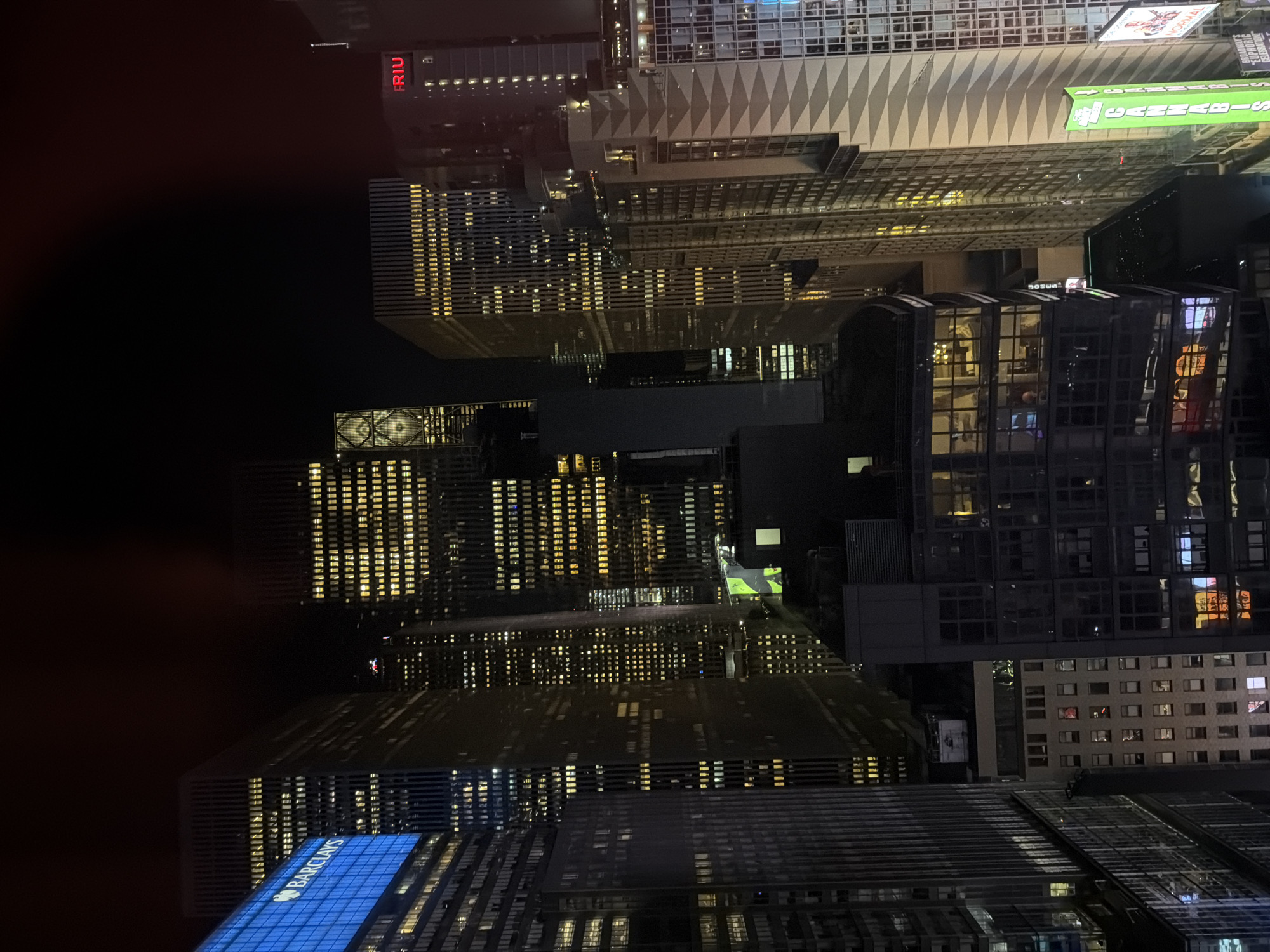 This media file captures a dense urban cityscape at night, viewed from a high vantage point looking down upon numerous skyscrapers and buildings. The dominant feature is the interplay of darkness and artificial light, with countless windows illuminated in warm yellow, cool white, and blue hues, creating a glowing grid pattern across the dark facades.
Many buildings are visible, showcasing various architectural styles. One prominent building in the upper central area features a distinctive diamond-patterned facade, while others are characterized by a multitude of rectangular windows. The deep black sky indicates it is late evening or night. No human figures or specific activities are discernible, but the illuminated windows suggest active offices or residences within the bustling city.
Visible text includes:
- **"RIU"** in red letters, displayed on a building towards the upper left.
- **"BARCLAYS"** in white lettering against a blue background, adorning the angled roofline of a building in the lower left.
- **"CANNABIS"** (or part of it) and **"NOOMA"** in green and white, respectively, are visible on signs attached to a building facade in the upper right quadrant, suggesting commercial establishments.
The scene conveys the vibrant, illuminated essence of a major metropolitan area after dark, consistent with a location like New York City. The artificial lights reflect off some surfaces, adding to the visual texture of the scene.
