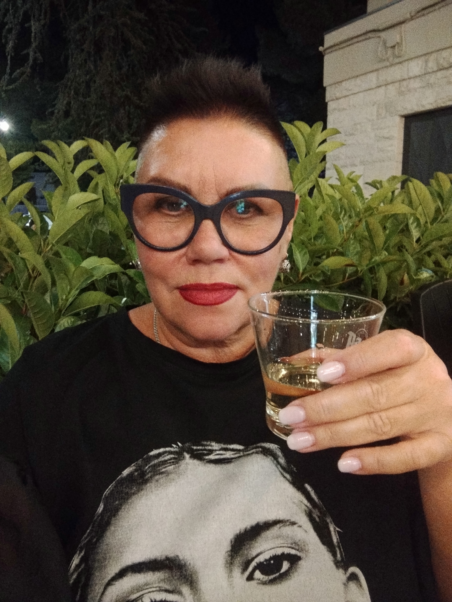 A middle-aged woman with dark hair styled upwards and wearing large dark-rimmed glasses and red lipstick holds a clear glass of amber liquid in her right hand. She is wearing a black t-shirt with a large grayscale image of a person's face printed on it. The woman is seated outdoors at night, with lush green foliage and a building in the background. The scene appears to be from a public space or restaurant patio in Toritto, Italy.