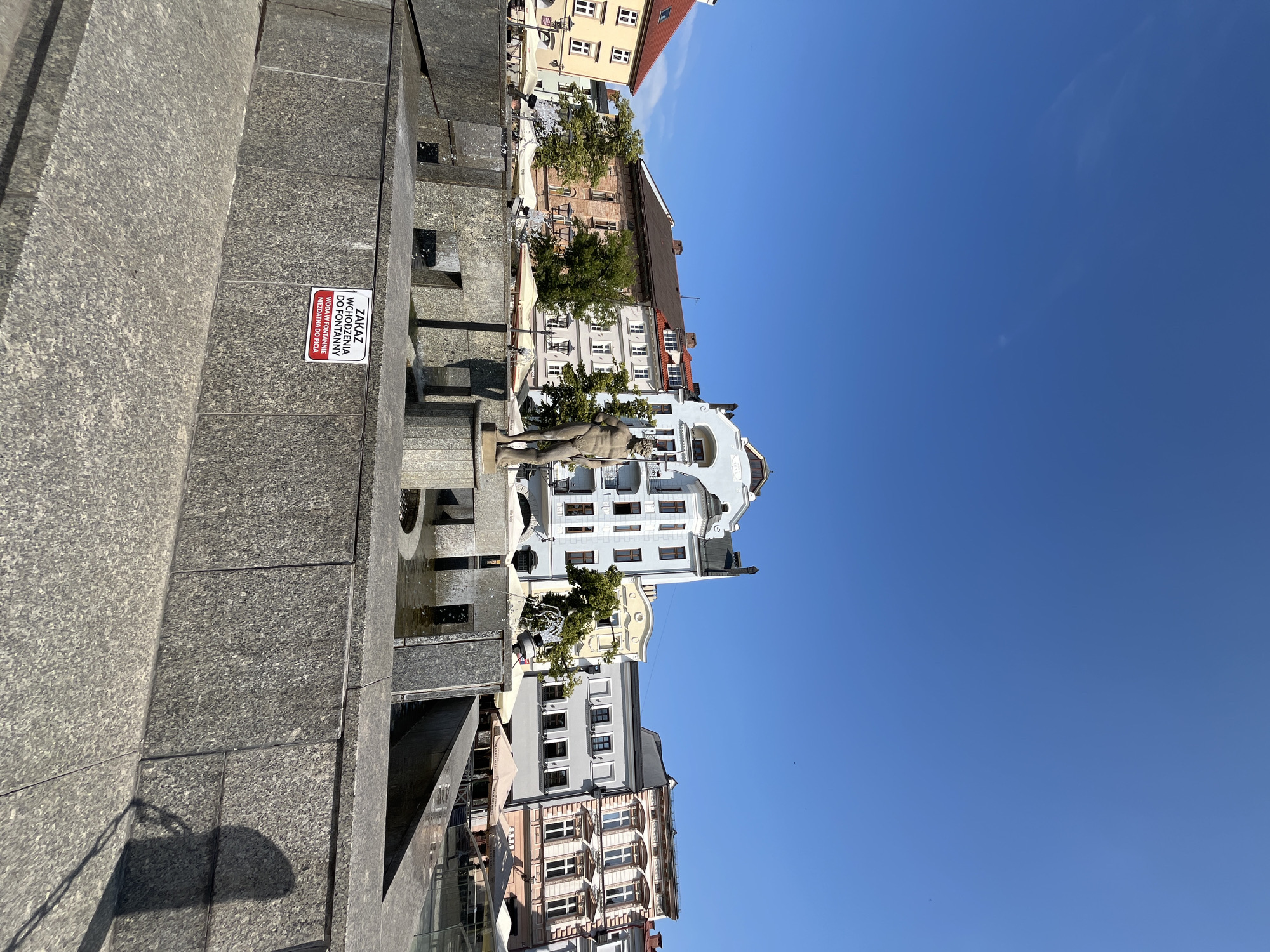 The original image shows a city square with a fountain in the foreground. A nude statue of a male figure stands in the center of the fountain. The statue is holding a trident. The fountain has a granite base with several water spouts. A sign on the side of the fountain reads "ZAKAZ WCHODZENIA DO FONTANNY WODA W FONTANNIE NIEZDATNA DO PICIA", which translates to "NO ENTERING THE FOUNTAIN WATER IN THE FOUNTAIN IS NOT SUITABLE FOR DRINKING". Behind the fountain, several multi-story buildings with various architectural styles line the square. The buildings are painted in different colors, including light blue, orange, and yellow. There are trees and umbrellas visible in front of the buildings. The sky is clear and blue. The overall scene appears to be a sunny day in a European city.