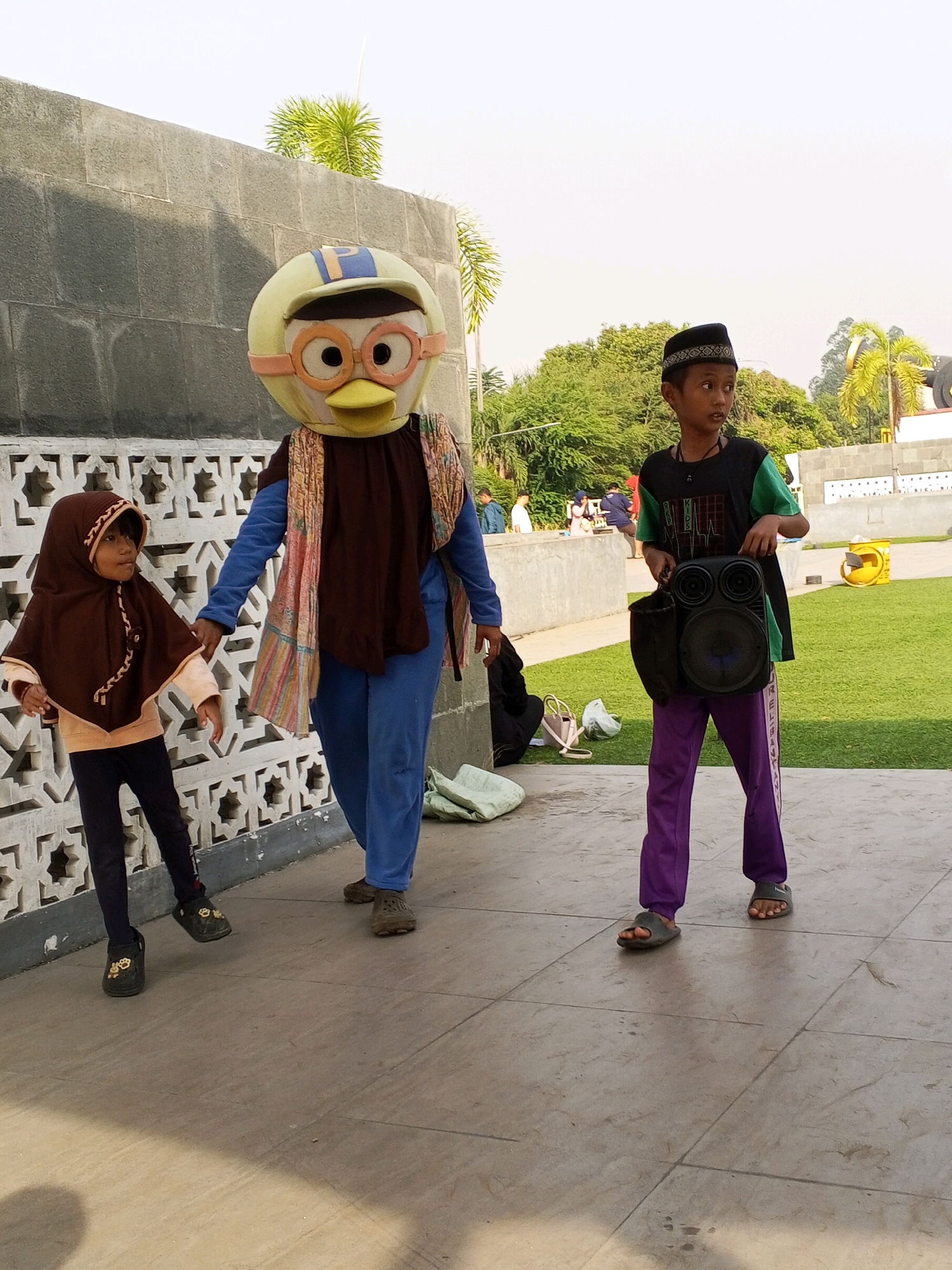 In Serang, Indonesia, a group walks outdoors on a paved area adjacent to a grassy lawn and a grey, textured wall. A person in a yellow duck costume with large goggles and an orange beak, wearing a blue jumpsuit and a colorful patterned scarf, walks with a child. The child, on the left, wears a brown hijab and a light brown top with navy leggings and black patterned shoes. On the right, a boy in a black and green t-shirt and purple track pants with white stripes, wearing a black kufi and grey sandals, carries a black portable speaker. Behind them, other individuals and a yellow trash bin are visible on the grass. The sky is overcast.