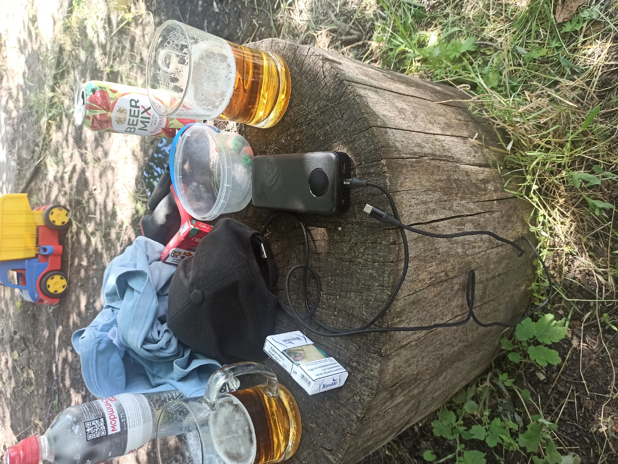 The image depicts an outdoor scene, likely in a park or wooded area, given the presence of a large log and grass. The setting suggests a casual gathering or a moment of relaxation. The time of day appears to be daytime, with natural sunlight casting shadows.
Several objects are scattered on and around the log. There are two beer mugs, one with a significant amount of beer and foam, and another empty one. A can of "BEER MIX SPECIAL BEER" with strawberry imagery is also present. A plastic container, possibly for food or snacks, sits nearby. A black baseball cap is placed on top of a light blue t-shirt.
A black power bank, branded with "hoco.", is connected to a charging cable, suggesting that someone is using or has used it to charge a device. Adjacent to the power bank is a pack of "Winston" cigarettes, with text in Cyrillic indicating "Quit smoking now" and other health warnings. A partially visible plastic bottle with a red cap, labeled "Моршинська" (a Ukrainian brand of mineral water), is also in the foreground. To the left, a yellow and red toy truck is visible.
The overall mood seems relaxed and informal, with the presence of beverages, cigarettes, and a comfortable setting. The items suggest a break or leisure activity, possibly a picnic or a stop during an outdoor excursion. The Ukrainian text on the water bottle and cigarette pack indicates the likely location of the scene to be Ukraine.