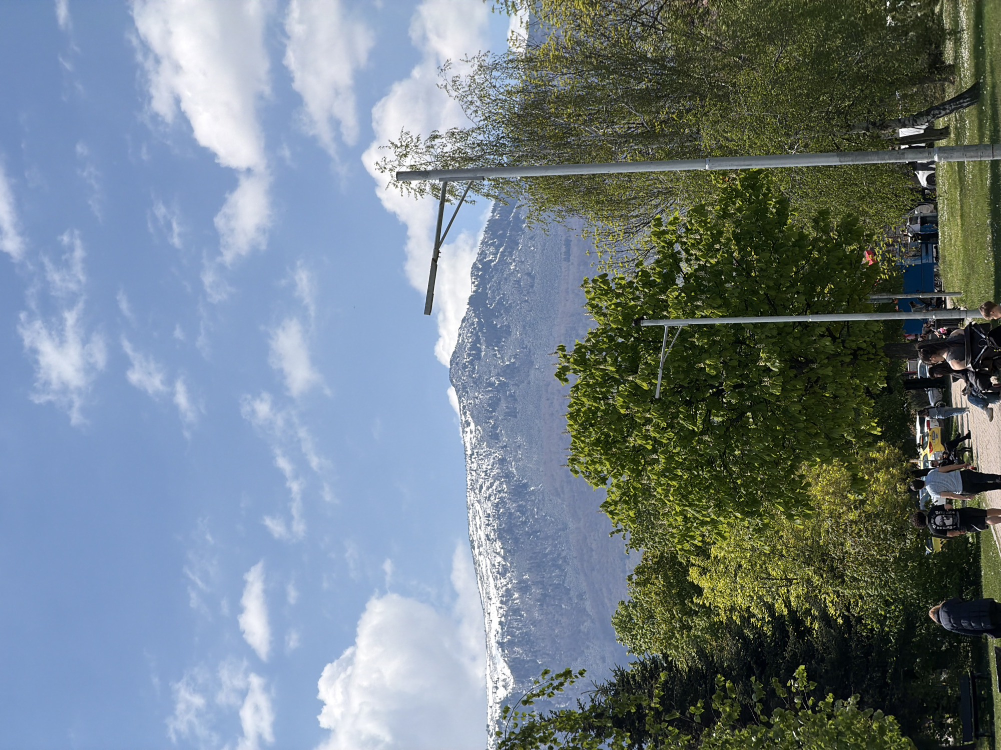 The image depicts a snow-capped mountain towering over a city park. The sky is blue with scattered white clouds. Trees with vibrant green foliage frame the mountain and line a park pathway. Several metal light posts are visible among the trees. A path with people walking and sitting runs along the edge of the park. The mountain is likely part of the Vitosha mountain range, which is visible from Sofia, Bulgaria. The park could be one of the city's many green spaces.