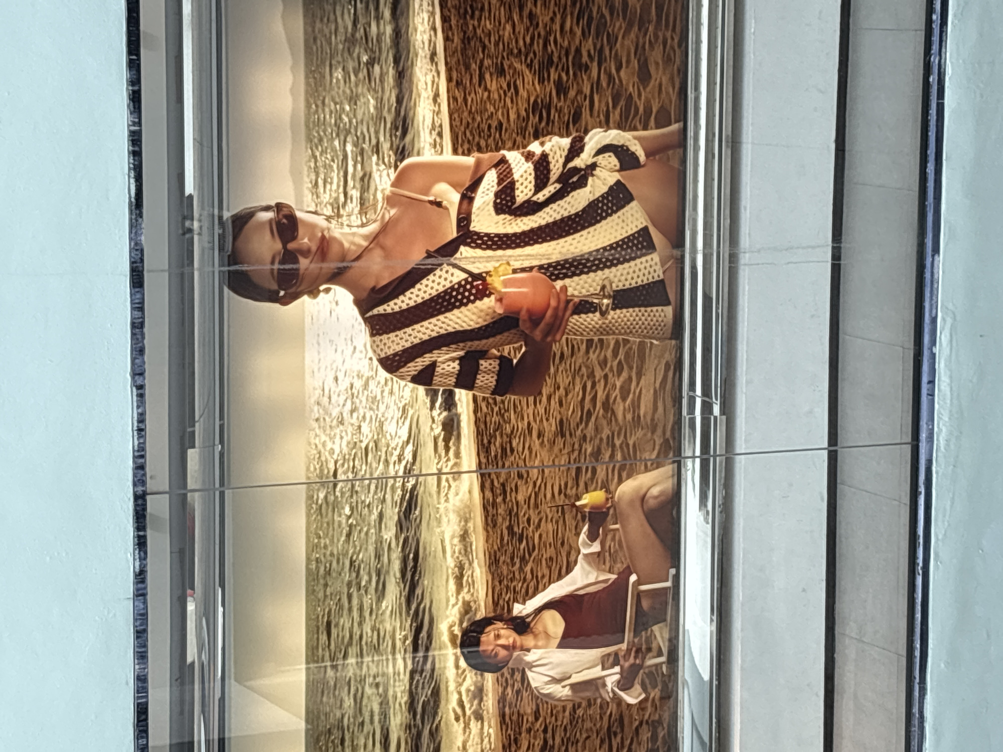 The image shows a reflection in glass of two different advertising images. The first image features a woman in sunglasses and a striped cover-up holding a cocktail in a beach setting. The second, reflected below, depicts another woman in a swimsuit and white cover-up sitting in a chair, also on a beach with a drink. The reflection suggests these advertisements are displayed in a location with tiled or mirrored surfaces. The location is identified as บ้านบางรักษ์, Thailand.
