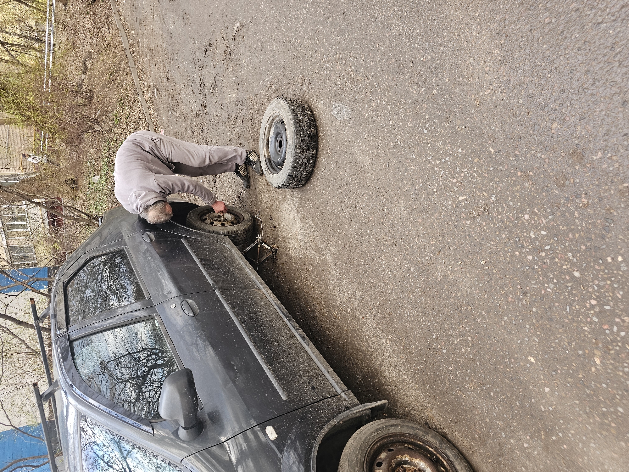 A man in a gray jumpsuit is changing a tire on a black car on a city street in Moscow, Russia. The car is parked on the side of the road, with a spare tire leaning against it. The man is bent over the car, working on the tire. A jack is visible under the car. The street is paved and appears to be in a residential area, with trees and buildings in the background. The day is overcast.