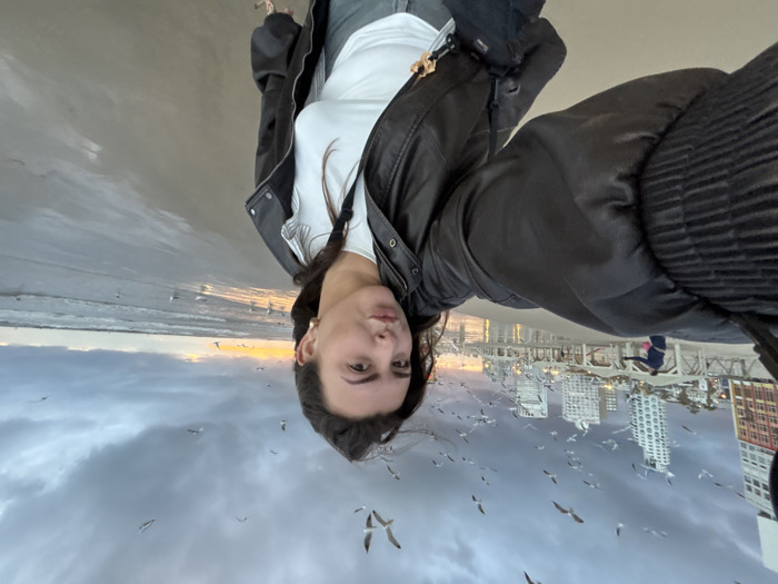 An upside-down selfie shows a woman on a beach against a cityscape. She wears a black jacket over a white shirt. The sky is cloudy with some birds flying. Buildings appear reflected in the water. Ocean waves are visible. The woman gazes into the camera.