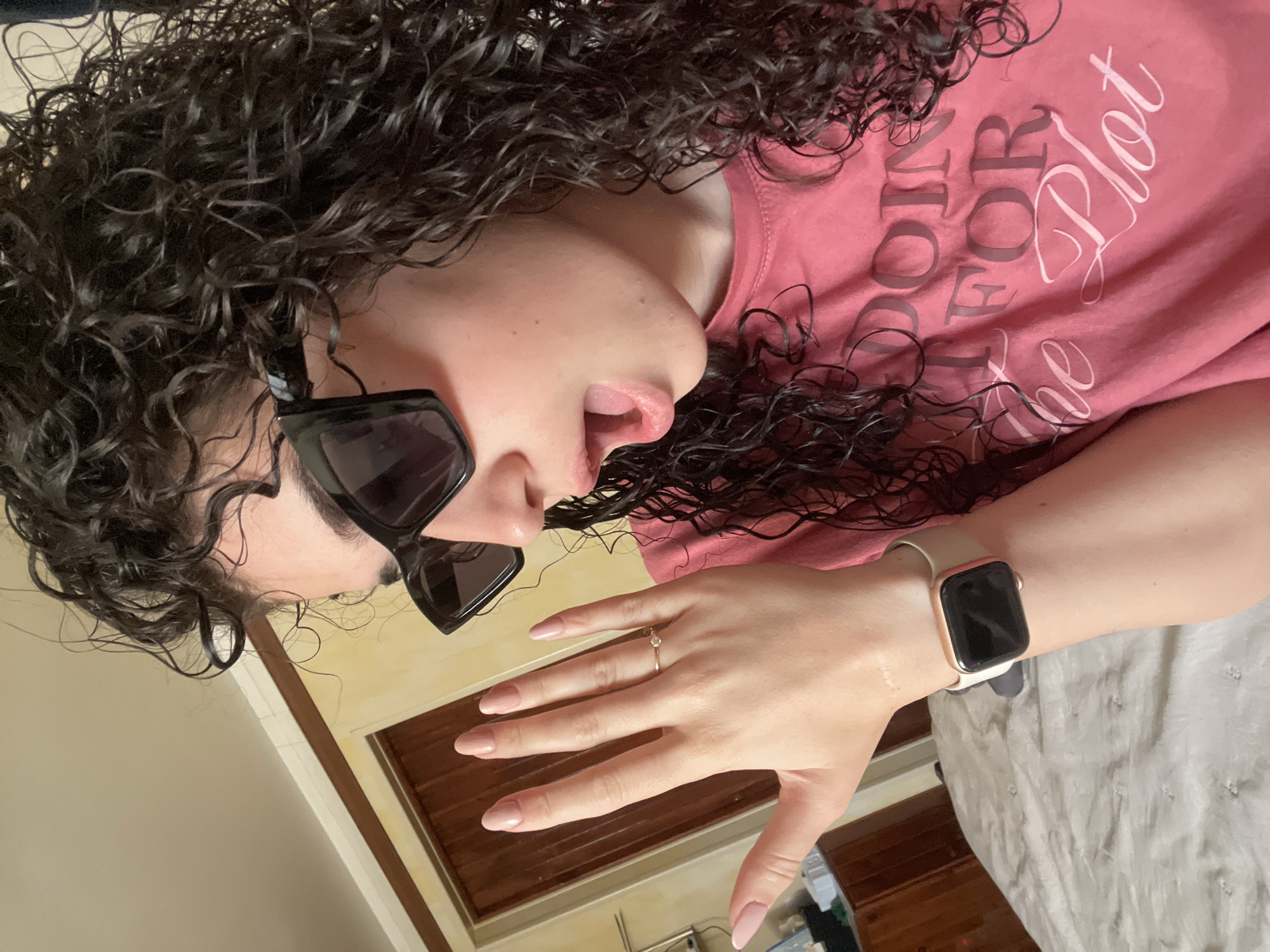 The image shows a person with curly dark hair, wearing sunglasses and a pink t-shirt with the words "Doing it for the Plot" printed on it. They are also wearing a ring on their ring finger and an Apple Watch on their wrist. The background appears to be an interior setting with wood trim and a beige wall. There is no indication of Sofia, Bulgaria in this image.