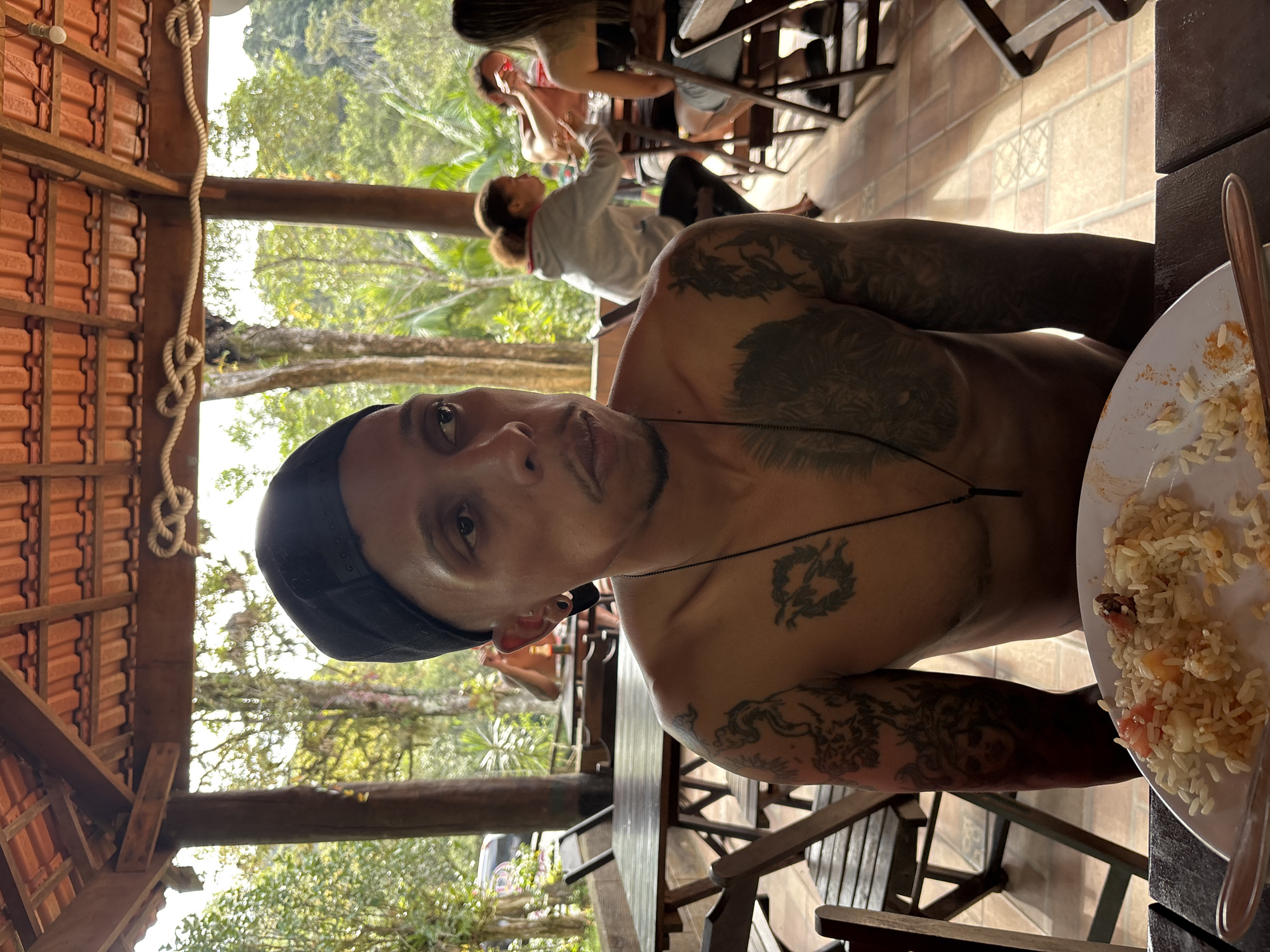 A medium, eye-level, full indoor/outdoor shot depicts a man, with his head and upper torso visible. He has light brown skin, dark eyes, and is wearing a black cap. He has a thin mustache and goatee. The man has visible tattoos on his chest and arms. A thin black necklace hangs around his neck. He is facing the camera.
To his right is a white plate filled with food. A fork is partially visible. Behind the man, other people are sitting at tables. The environment is open, with visible trees and foliage in the background. The structure overhead appears to be a covered patio or open-air restaurant, with wooden beams and a tiled roof.
There is no location information to suggest the city of São Paulo, Brazil.
