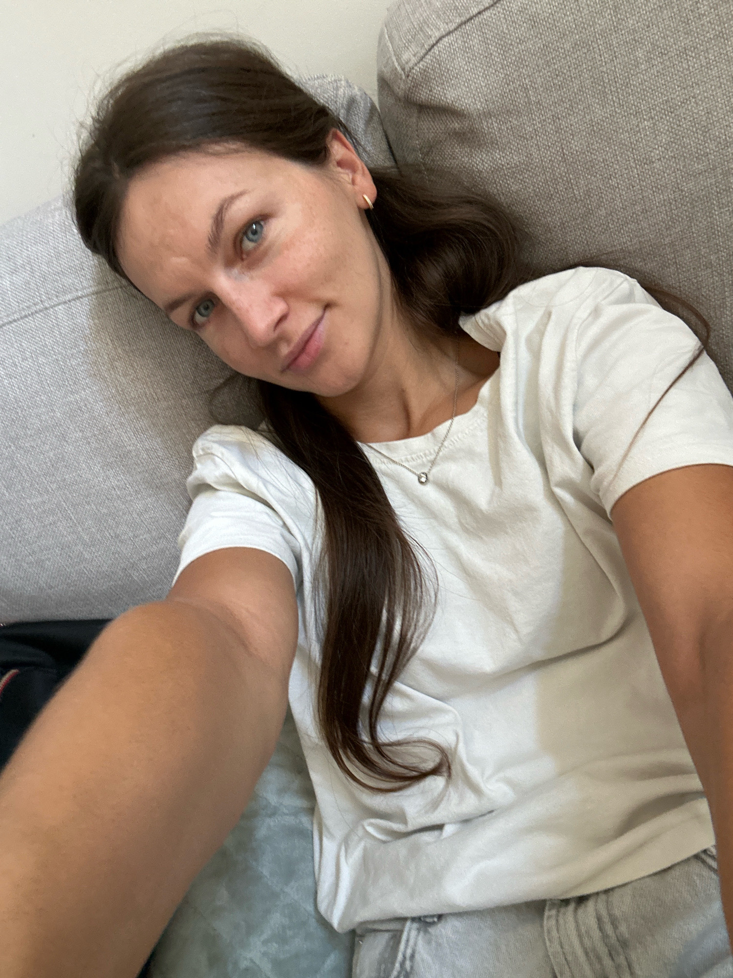 A young woman with long, dark hair and blue eyes is lying on a sofa, taking a selfie. She is wearing a white t-shirt and grey shorts, and a small silver necklace. Her left arm is extended towards the camera, and her right arm is resting on the sofa. The background shows a textured grey sofa and a blue-green patterned blanket. The lighting is natural, suggesting it is daytime. The location is a residential interior, possibly in Boreti, Montenegro.