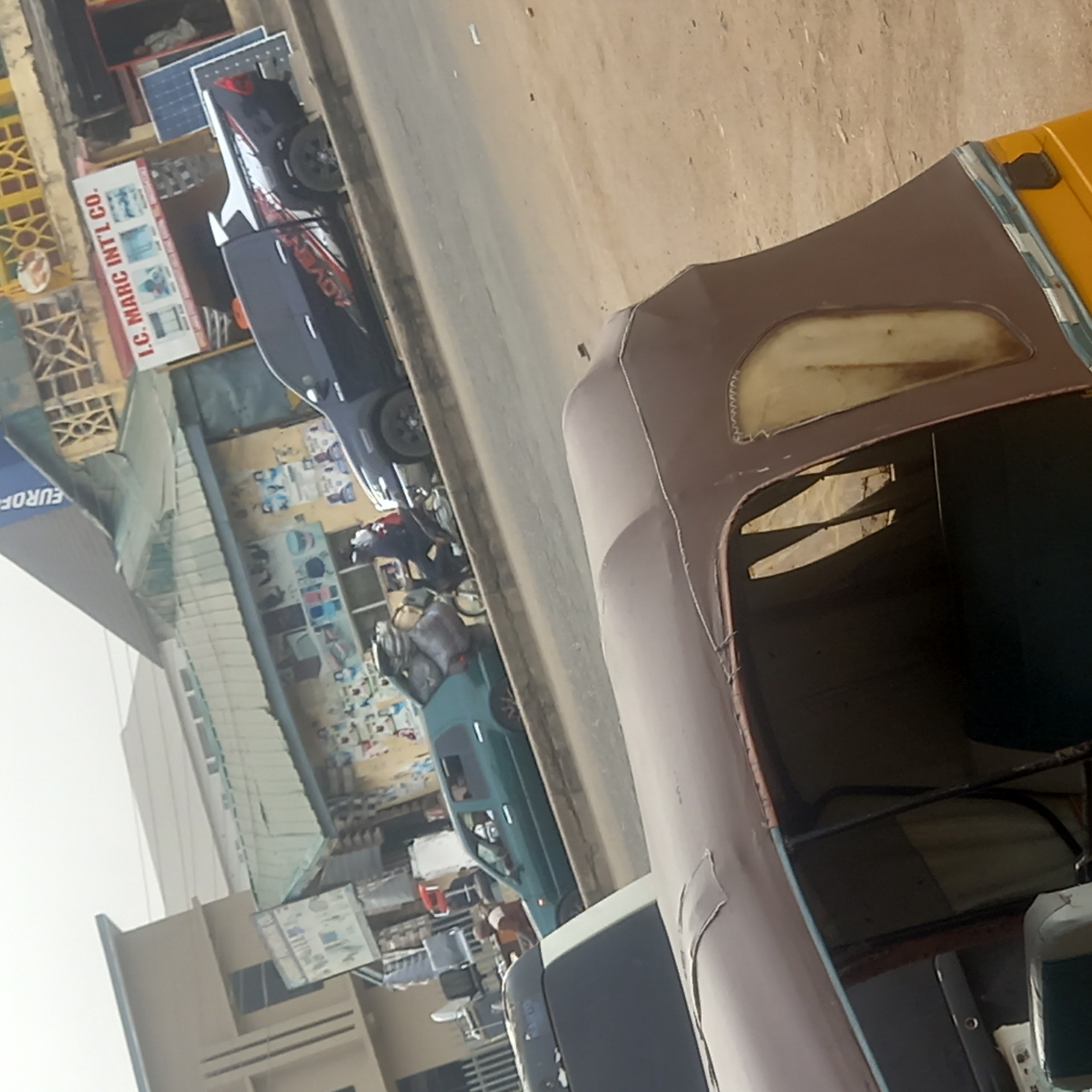 This media file captures a busy daytime street scene, likely in a commercial area of Dadi Riba, Nigeria. The image is tilted to the right, presenting the street and its adjacent buildings from a slightly elevated perspective.
In the immediate foreground, a significant portion of a light brown or beige auto-rickshaw, commonly known as a Keke Napep, dominates the right side. Its canvas roof and part of its interior are visible, showing worn seats and a pattern that partially reads "WWW.AN".
The midground features a paved road running parallel to the foreground vehicle. Along the left side of the street (as viewed in the tilted image), several buildings house various businesses. A prominent white sign clearly displays "L.C. MARC INT'L CO." with images of what appear to be air conditioning units or other electrical appliances. Near this sign, a dark blue car and a dark-colored motorcycle, with "ADVENT" partially visible on its side, are parked. Behind them, several blue solar panels are visible, possibly on display or installed. Further along the street, a green car is parked, and a person can be faintly seen inside. Another partial sign with "EUROF" (likely part of "EUROPE") is also visible. The walls of some buildings are covered with numerous posters and papers, suggesting a vibrant, active environment.
The scene depicts typical daily street activity under bright daylight, implying sunny weather. The types of vehicles, commercial establishments, and general urban setting are characteristic of an urban or semi-urban area in Nigeria.