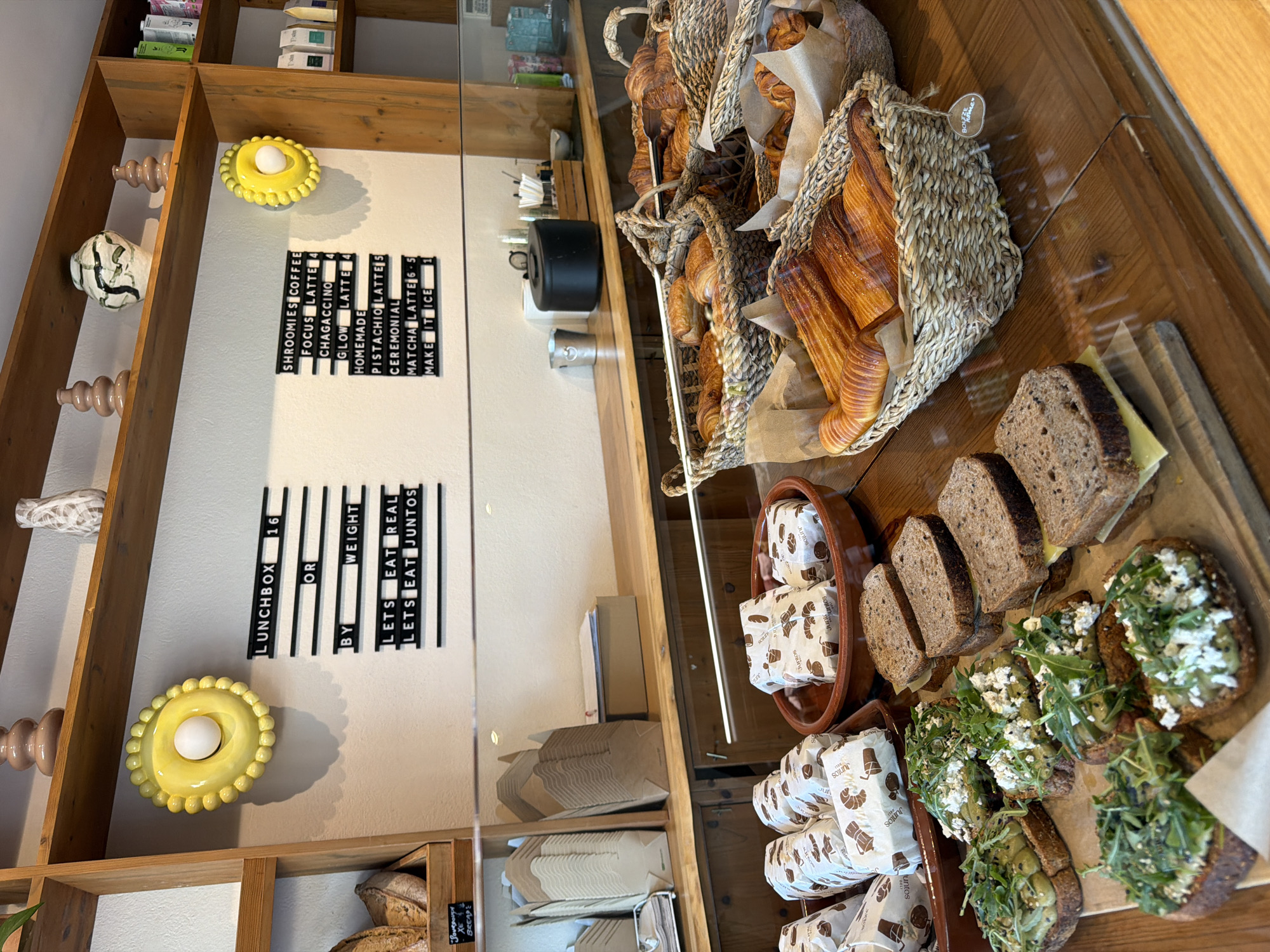 This image depicts the interior of a cafe in Santa Eulària des Riu, Spain. The scene is viewed from a slightly elevated angle, looking down onto a wooden counter displaying various food items and cafe signage.
On the counter, in the foreground, are several open-faced sandwiches on dark bread, topped with avocado, cheese, and greens. Behind these, a terracotta dish holds individually wrapped pastries or sandwiches. Further back, a large wicker basket overflows with golden-brown croissants. Another basket next to it contains more pastries. To the left, a stack of disposable brown paper containers is visible.
On the wall behind the counter, two black letter boards are mounted. The left board displays "LUNCHBOX 16 OR BY WEIGHT LETS EAT REAL LETS EAT JUNTOS." The right board lists menu items and prices: "SHROOMIES COFFEE 4 FOCUS LATTE 4 CHAGACCINO 4 GLOW LATTE 4 HOMEMADE PISTACHIO LATTE 5 CEREMONIAL MATCHA LATTE 6.5 MAKE IT ICE 1."
Decorative elements on a wooden shelf above the letter boards include two ribbed brown ceramic vases and a white and green patterned vase. A bright yellow, circular wall decoration with a white central element is positioned to the right of the "LUNCHBOX" sign. Further to the right on the shelves are small packaged items, possibly food or drink.
The environment appears clean and well-lit, suggesting a commercial establishment. No people are visible in the frame. The overall impression is of a welcoming cafe with a focus on baked goods and light meals.