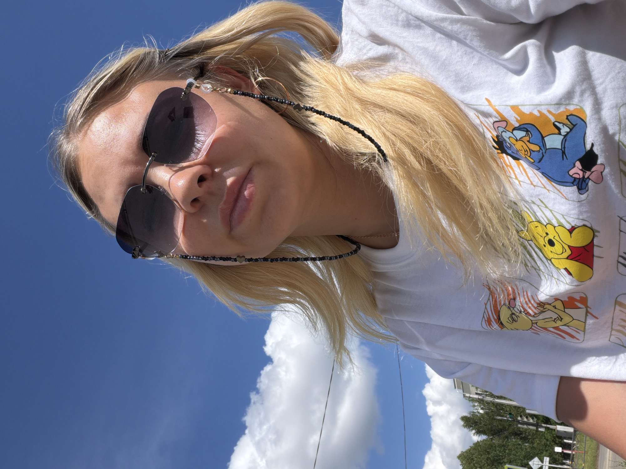 The media file is a close-up, low-angle shot of a young blonde woman outdoors on a sunny day. She is wearing sunglasses and a white t-shirt featuring cartoon characters, possibly from Winnie the Pooh. A black beaded string is attached to her sunglasses and loops around her ear, with a small metallic heart charm. The background consists of a bright blue sky with scattered white clouds, indicating fair weather. Sunlight illuminates the woman's face and hair, suggesting it is daytime. The tilt of the image and the upward perspective imply the photo was taken looking up at the sky. Visible text includes cartoon graphics on the t-shirt. The location context is Chaikovsky, Russia, though specific landmarks are not clearly identifiable in this close-up. The woman's expression is neutral.
