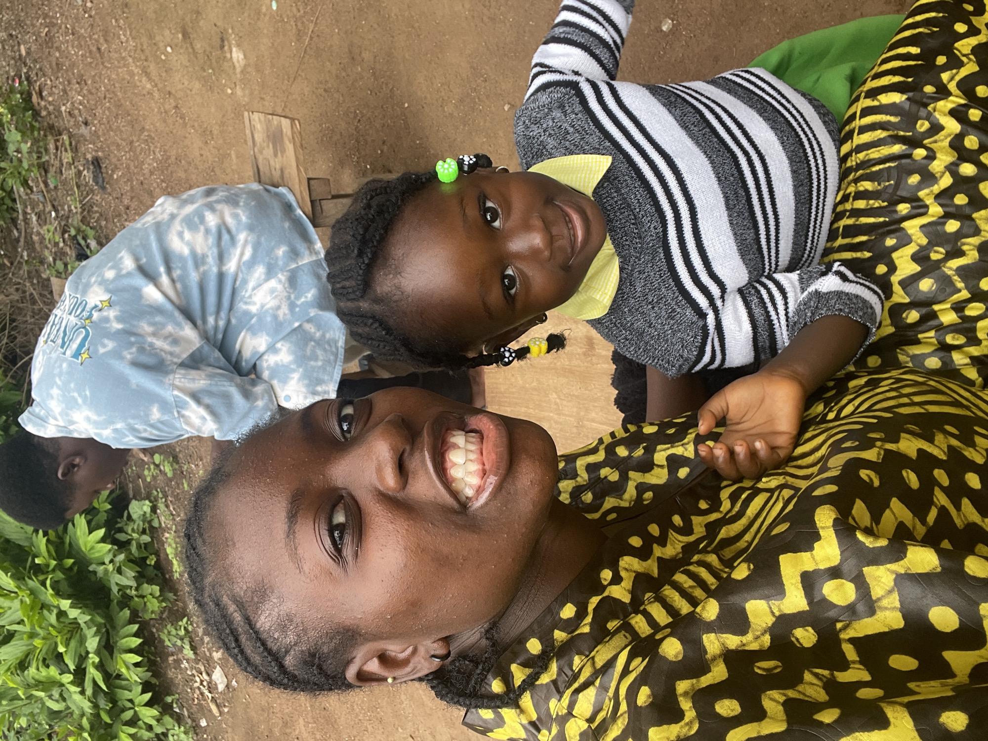 In Akwanga, Nigeria, an outdoor scene unfolds with three individuals. In the foreground, a young woman with braided hair smiles broadly at the camera, her mouth open to reveal her teeth. She wears a vibrant yellow and black patterned garment. Beside her, a young child with braided hair, adorned with colorful beads, looks towards the camera with a gentle smile. The child is dressed in a grey and white striped sweater with a yellow collar and a green skirt. In the background, a third person, partially visible, is seated on a wooden bench and wearing a light blue tie-dye shirt. The ground appears to be reddish-brown earth, with patches of green foliage visible behind the individuals. The setting suggests a casual, communal outdoor space.