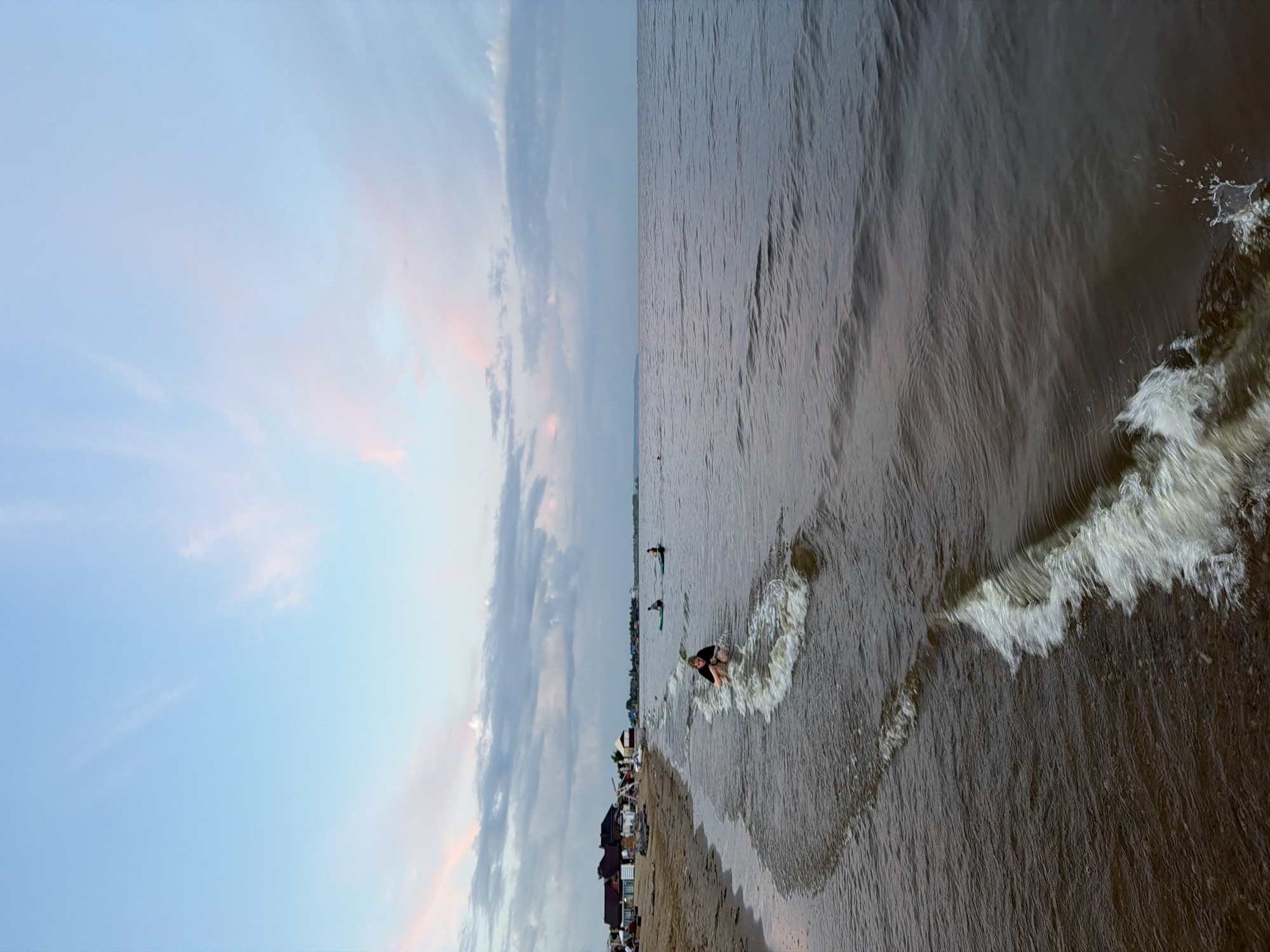 This image depicts a serene scene on the shores of the Kama River in Laishevo, Russia. The sky is a soft blend of pale blue and pink, with wispy clouds hinting at the setting sun. The gentle waves of the river lap at the sandy beach, where a young person is crouched in the shallow water, seemingly enjoying the cool embrace of the river. Further out in the water, two individuals are seen on paddleboards, navigating the calm surface. The background reveals a stretch of beach lined with small buildings, likely residences or small businesses, adding a touch of human presence to the natural landscape. The overall atmosphere is tranquil, capturing a moment of peaceful recreation by the river.