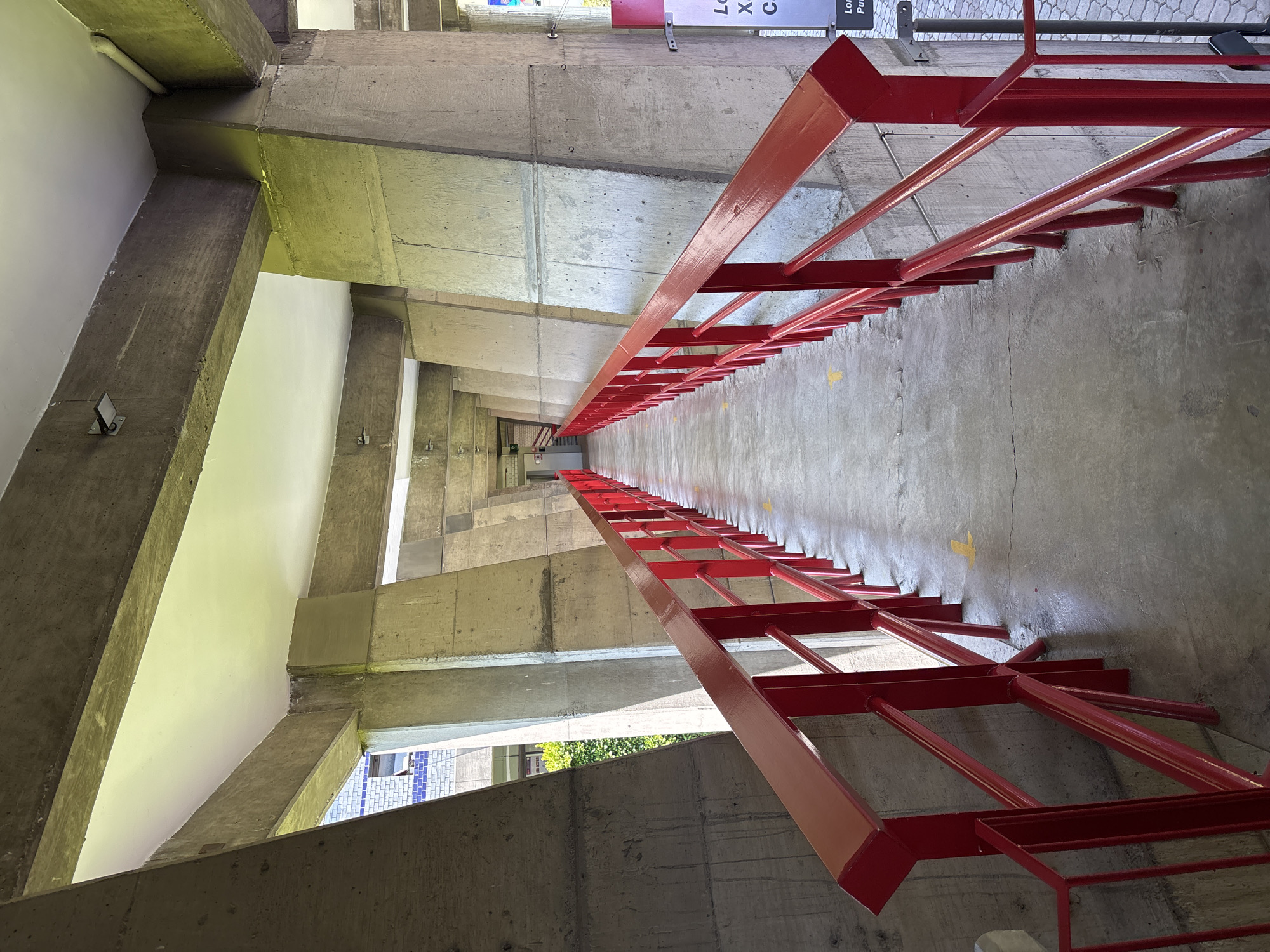 The image shows a long concrete staircase with a red metal railing in San Bernardino Tlaxcalancingo, Mexico. The walls are made of concrete and painted white in some sections. The staircase is angled upwards and appears to be outdoors. Yellow arrows are painted on the concrete steps. The photo is taken from a low angle looking up the staircase.