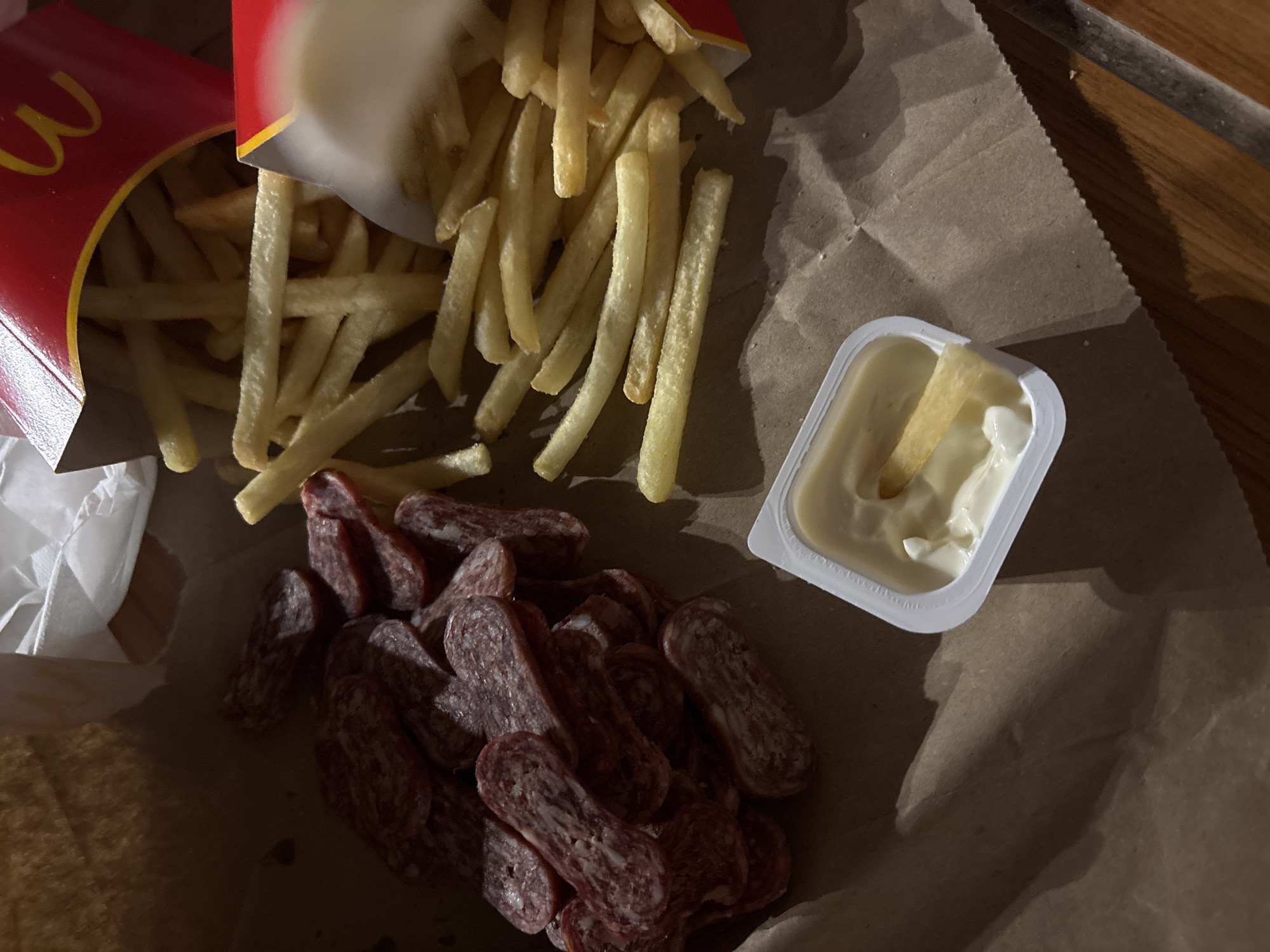 The image shows a close-up, overhead shot of a meal consisting of McDonald's french fries, salami slices, and a container of sauce. The fries are spilling out of two McDonald's cartons onto a brown paper surface. A white plastic container holds a light-colored sauce, with a single fry partially submerged in it. Below the fries and to the left, a pile of sliced salami is visible. The image is dimly lit, with shadows cast across the scene. No people are visible. The location is not specified, and there is no information to suggest that it is Plovdiv, Bulgaria.